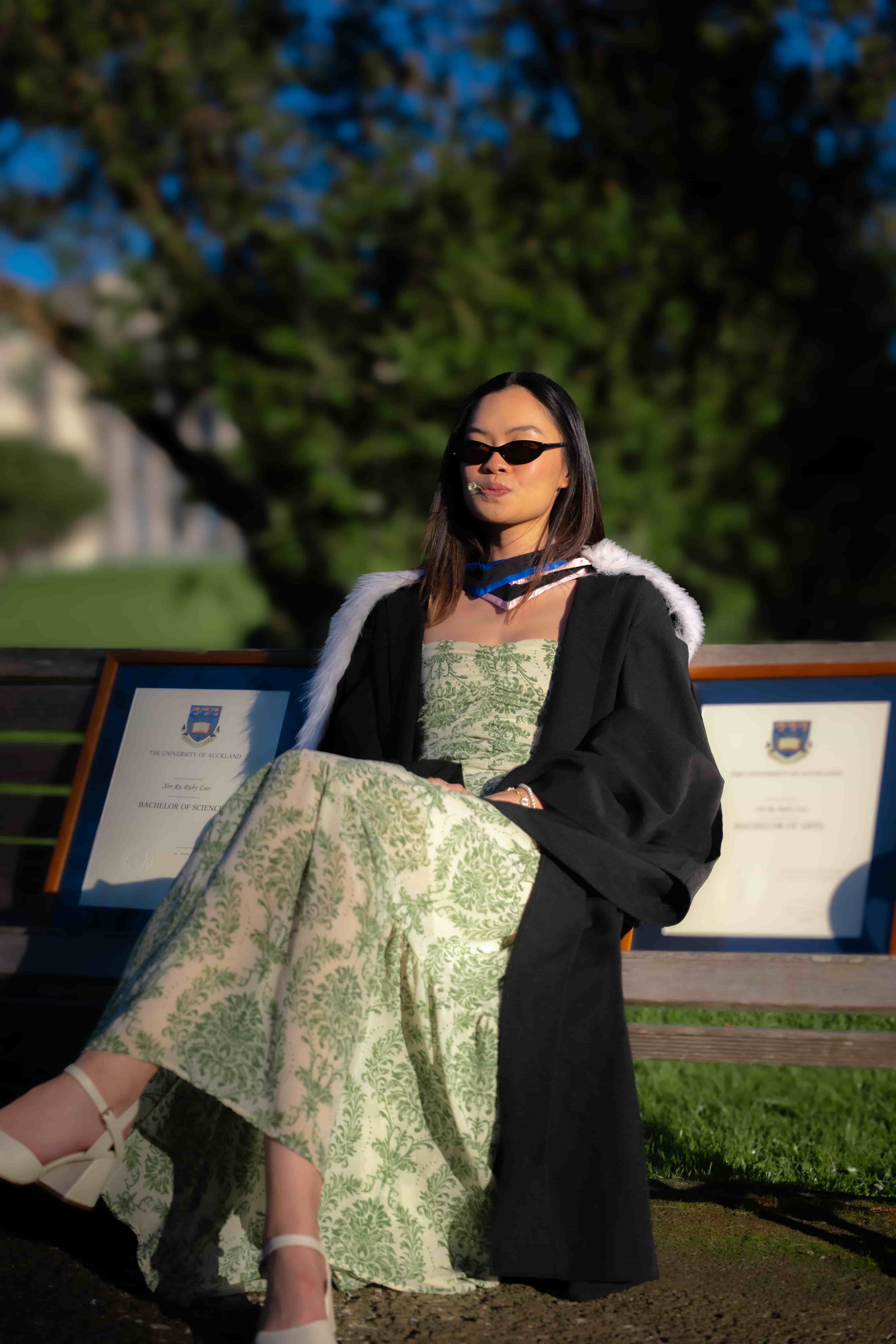 Ruby sitting on a park bench in sunglasses with her framed degrees on either side