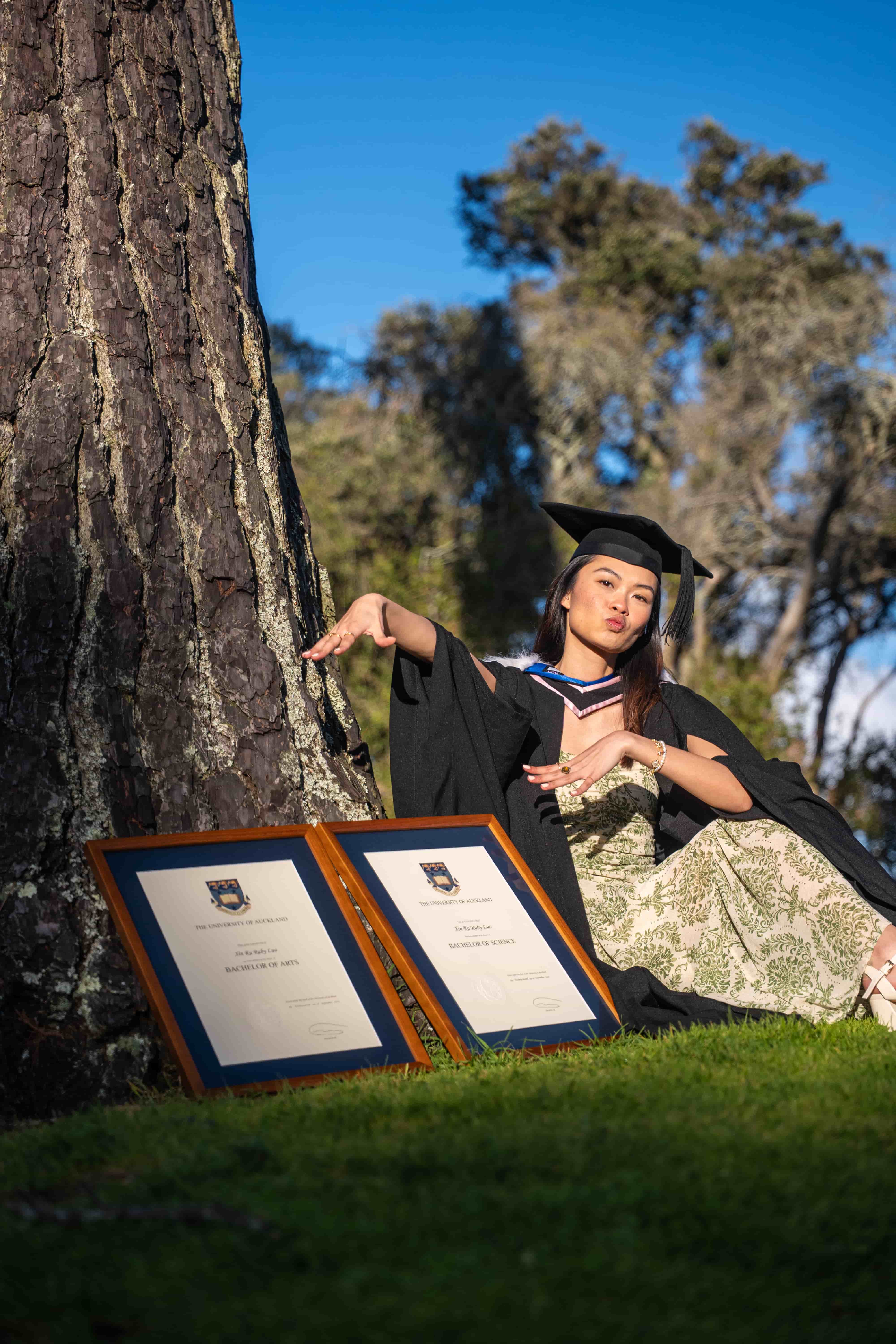Ruby striking a playful pose under a pine tree beside her framed degrees in golden light