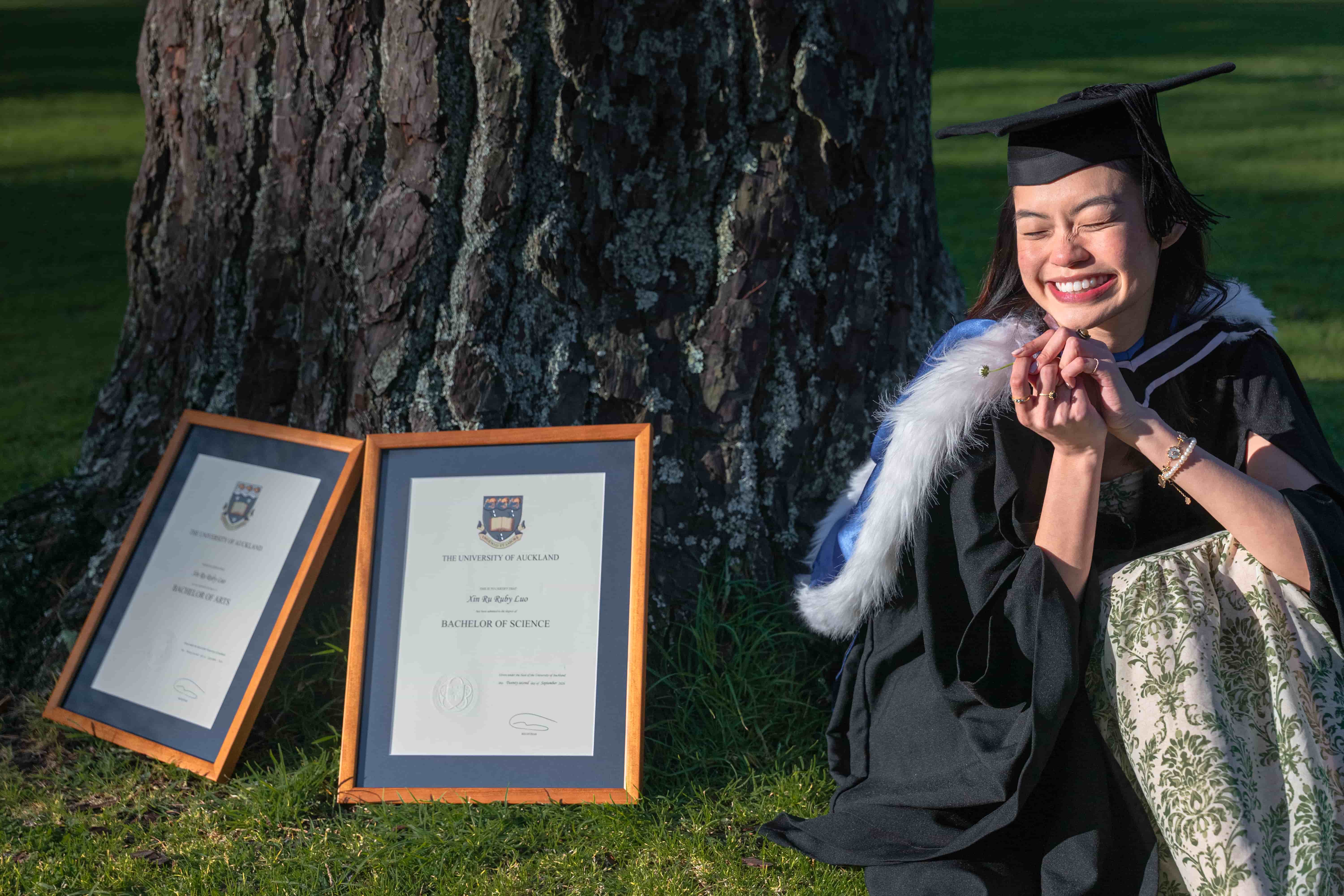 Ruby sitting on the grass beside a pine tree with her framed degrees leaning against it