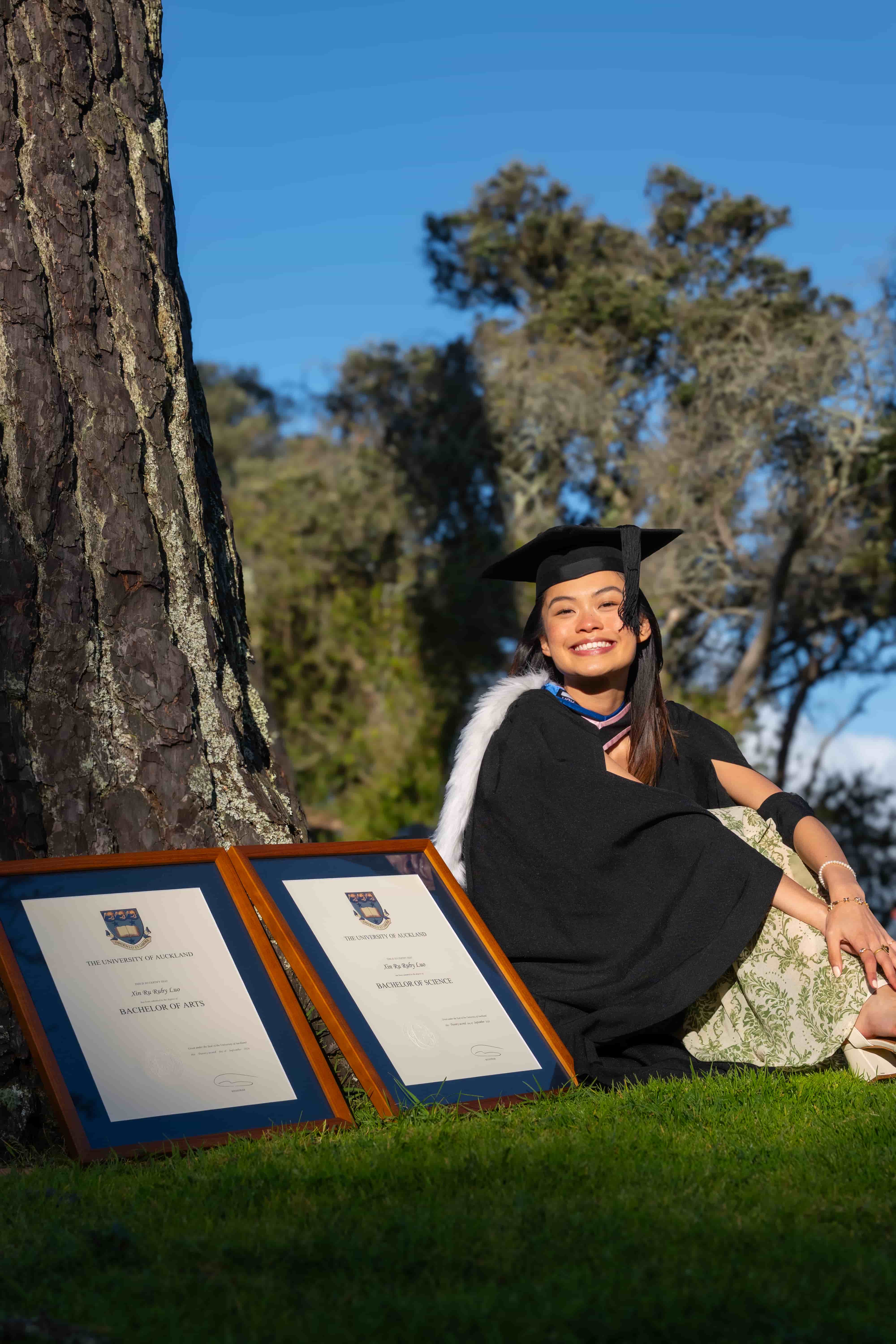 Ruby smiling on the grass under a pine tree with her University of Auckland degrees