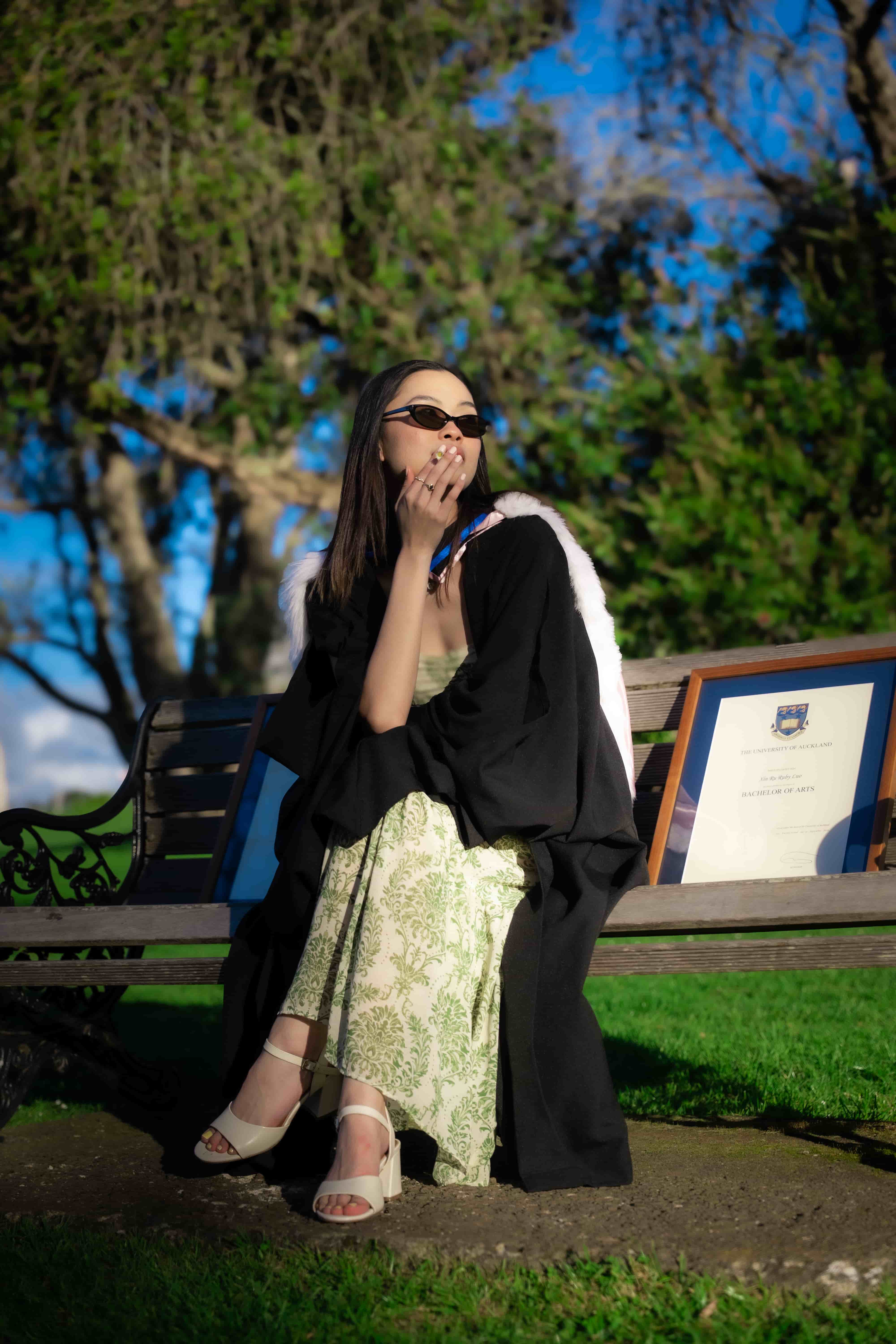 Ruby seated on a bench in the Auckland Domain wearing her graduation gown and green floral dress