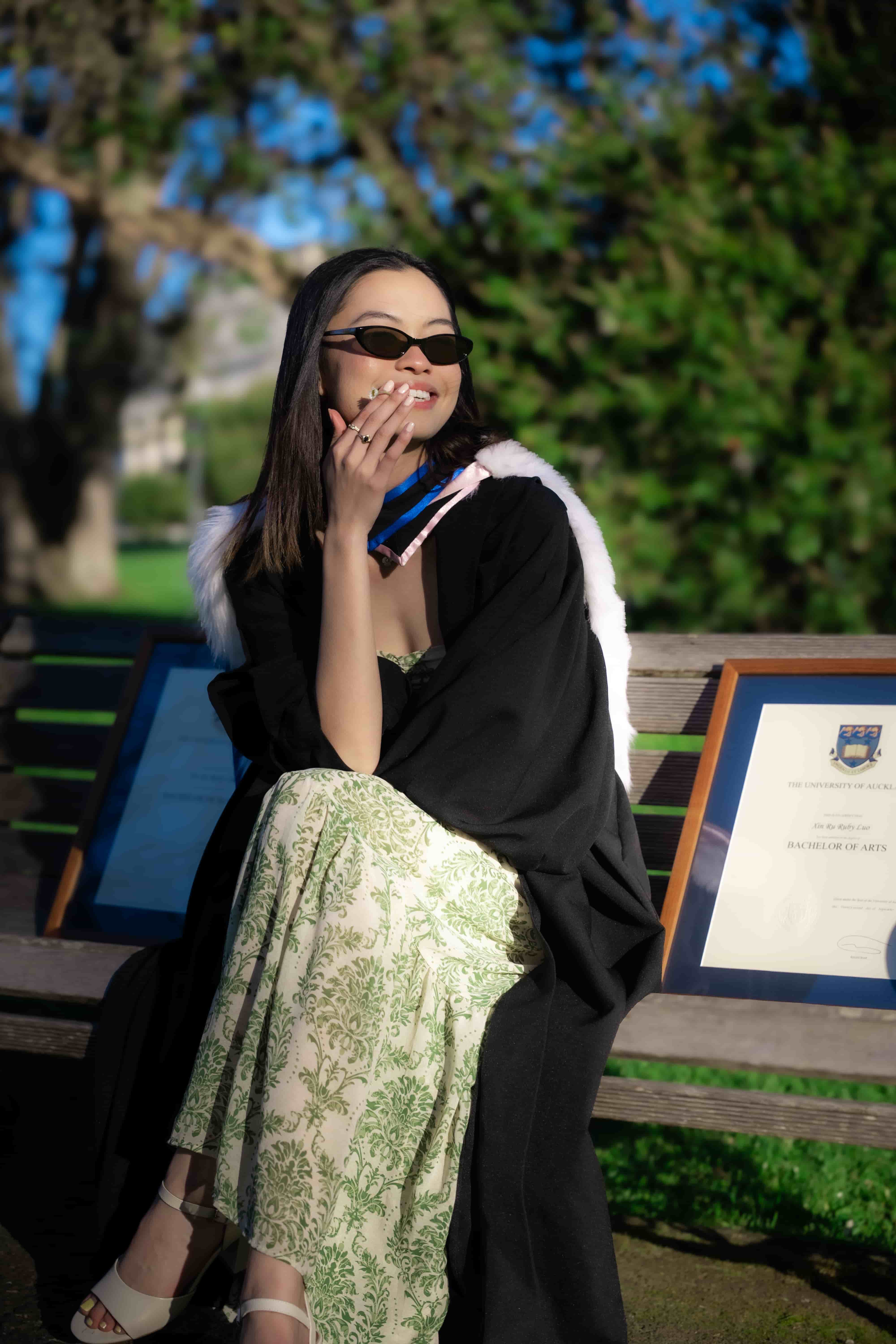 Ruby laughing in sunglasses on a park bench with her degrees and fur-trimmed graduation hood