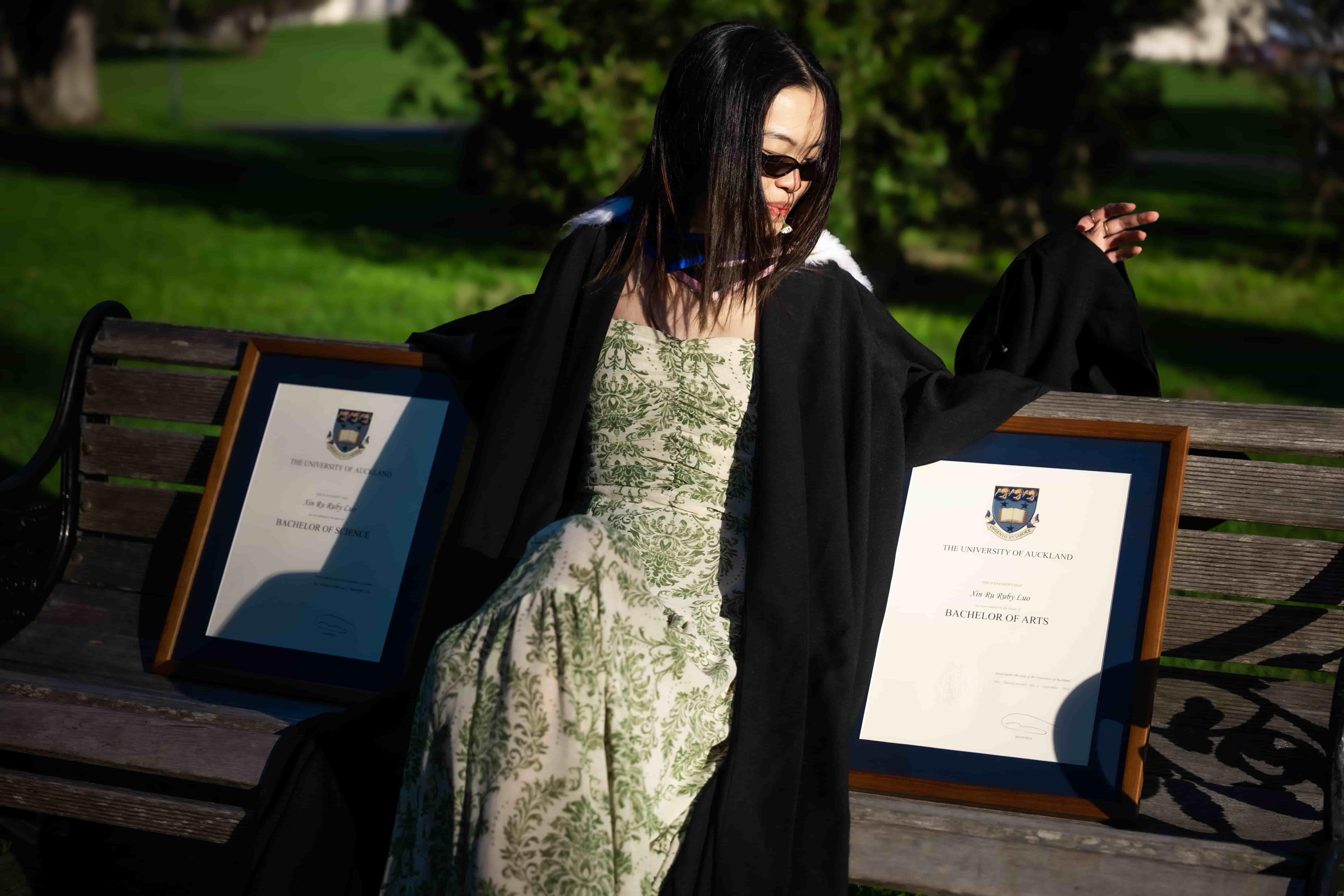 Ruby in sunglasses posing on a park bench between her two framed degrees