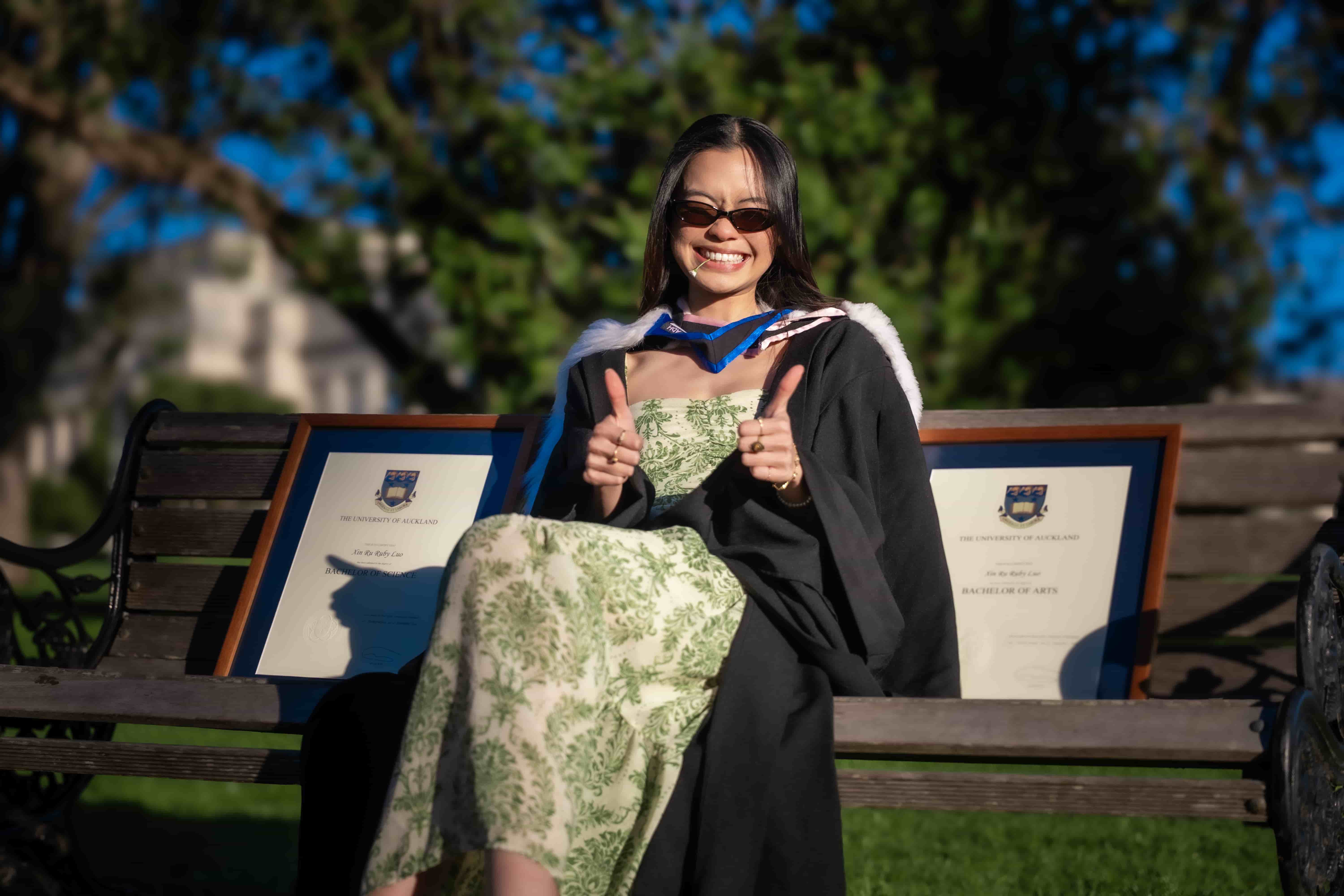 Ruby giving two thumbs up on a park bench between her framed degrees