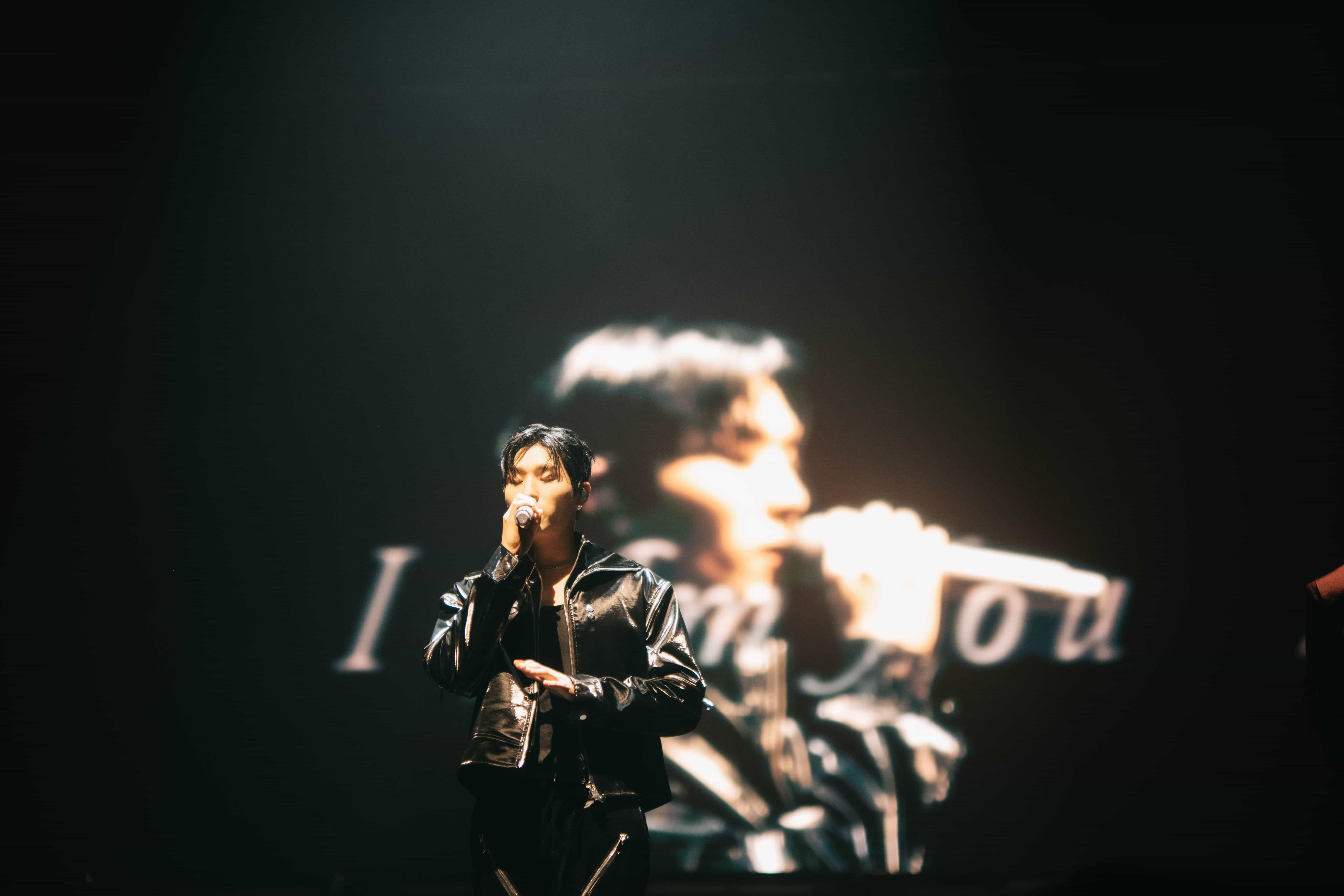 P1Harmony member in a leather jacket singing before a screen showing his close-up and lyrics