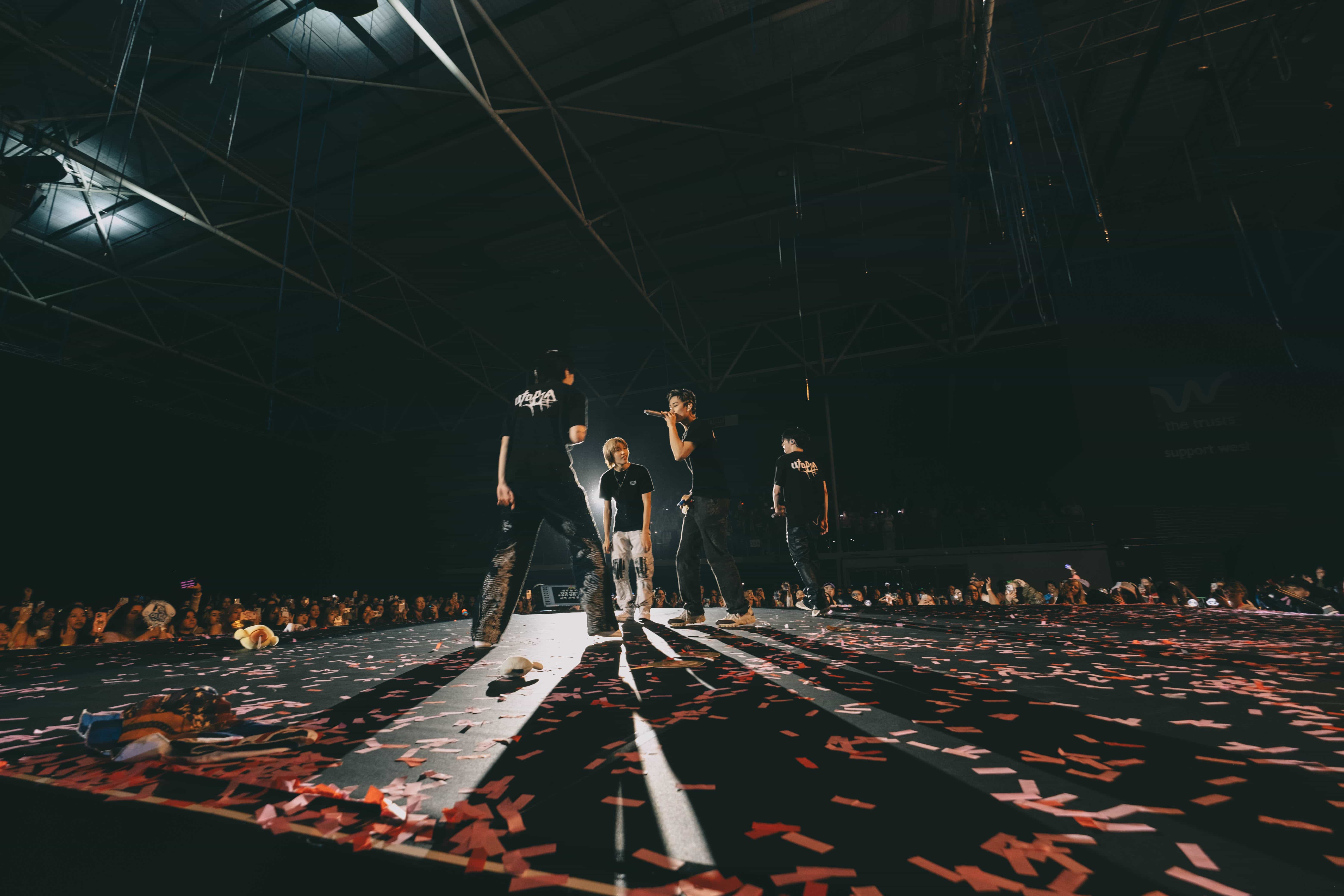 Wide view of P1Harmony members walking a long runway scattered with red confetti and light beams