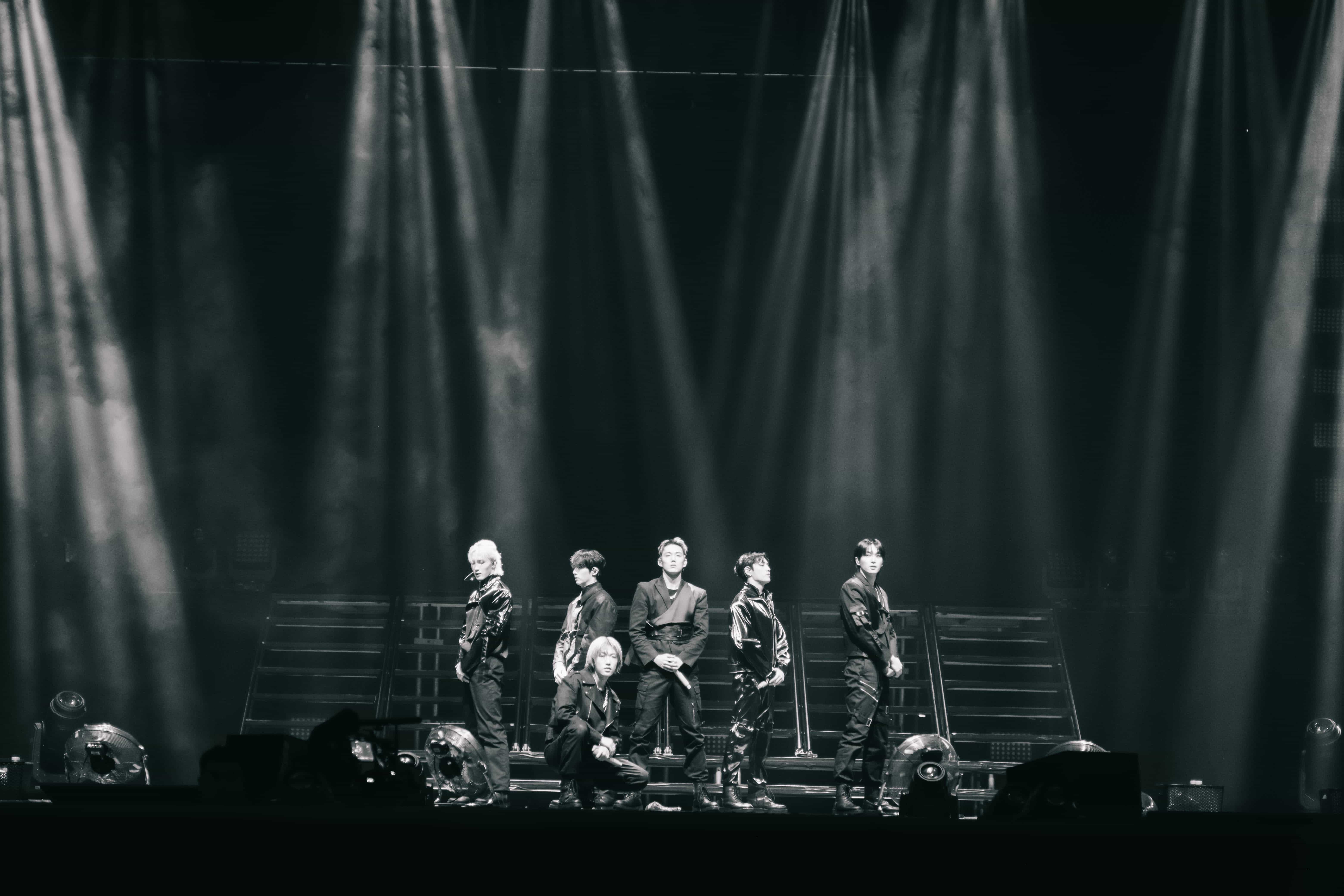 Black and white view of six P1Harmony members posed on stage stairs under sweeping spotlight beams