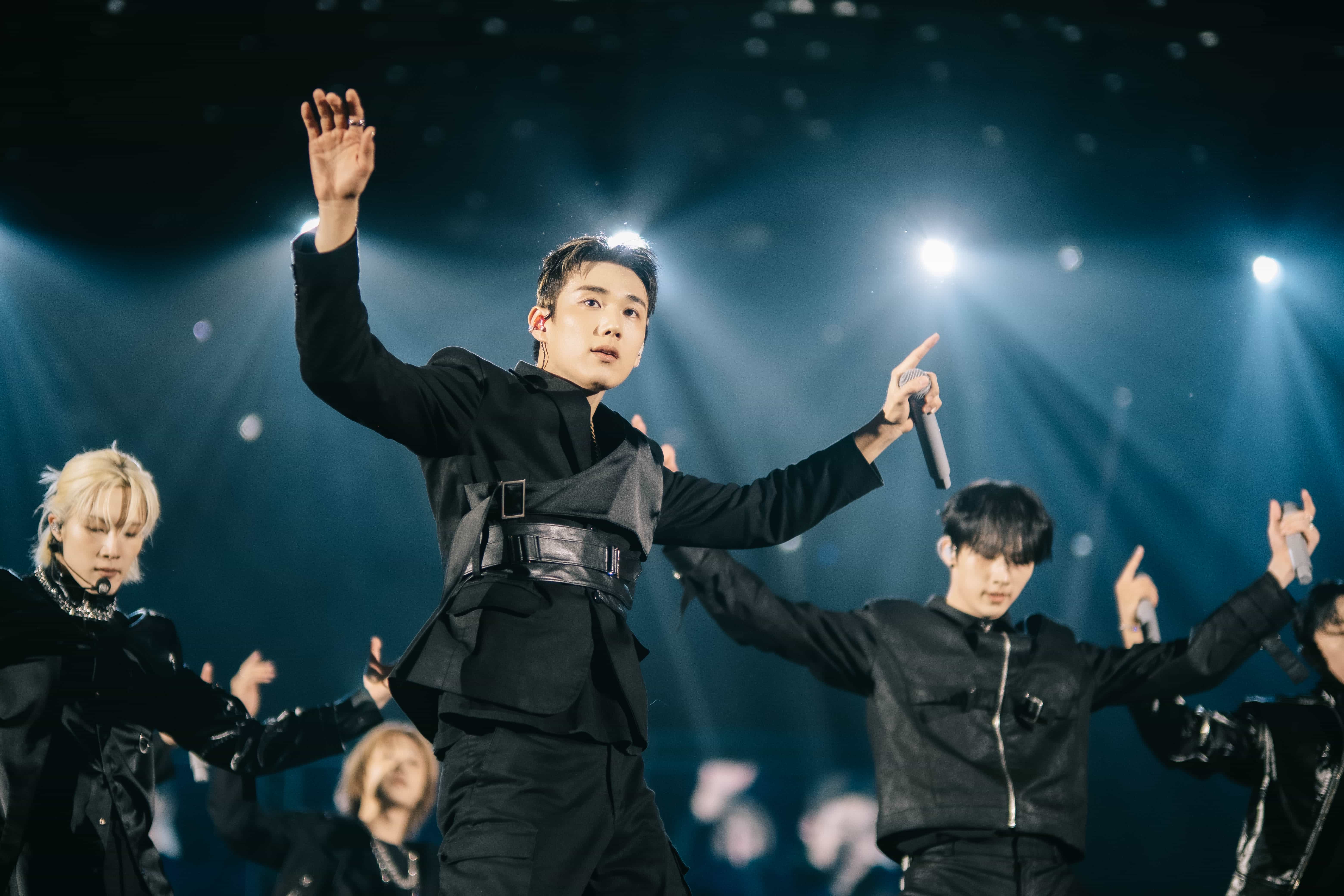 P1Harmony member in a black harness jacket raising one arm mid-dance, bandmates in step behind