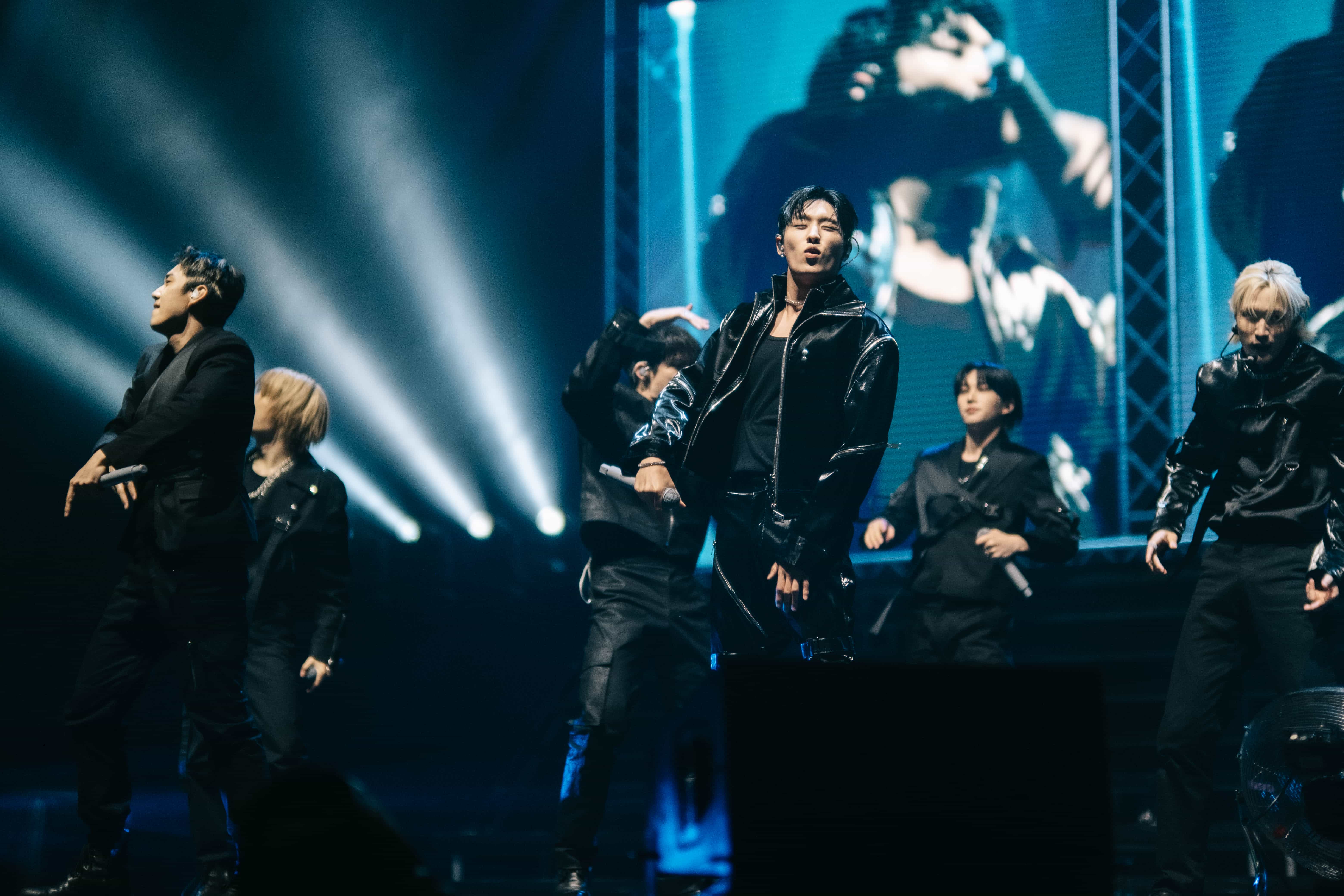 P1Harmony members in black leather dancing under white light beams with a live screen behind