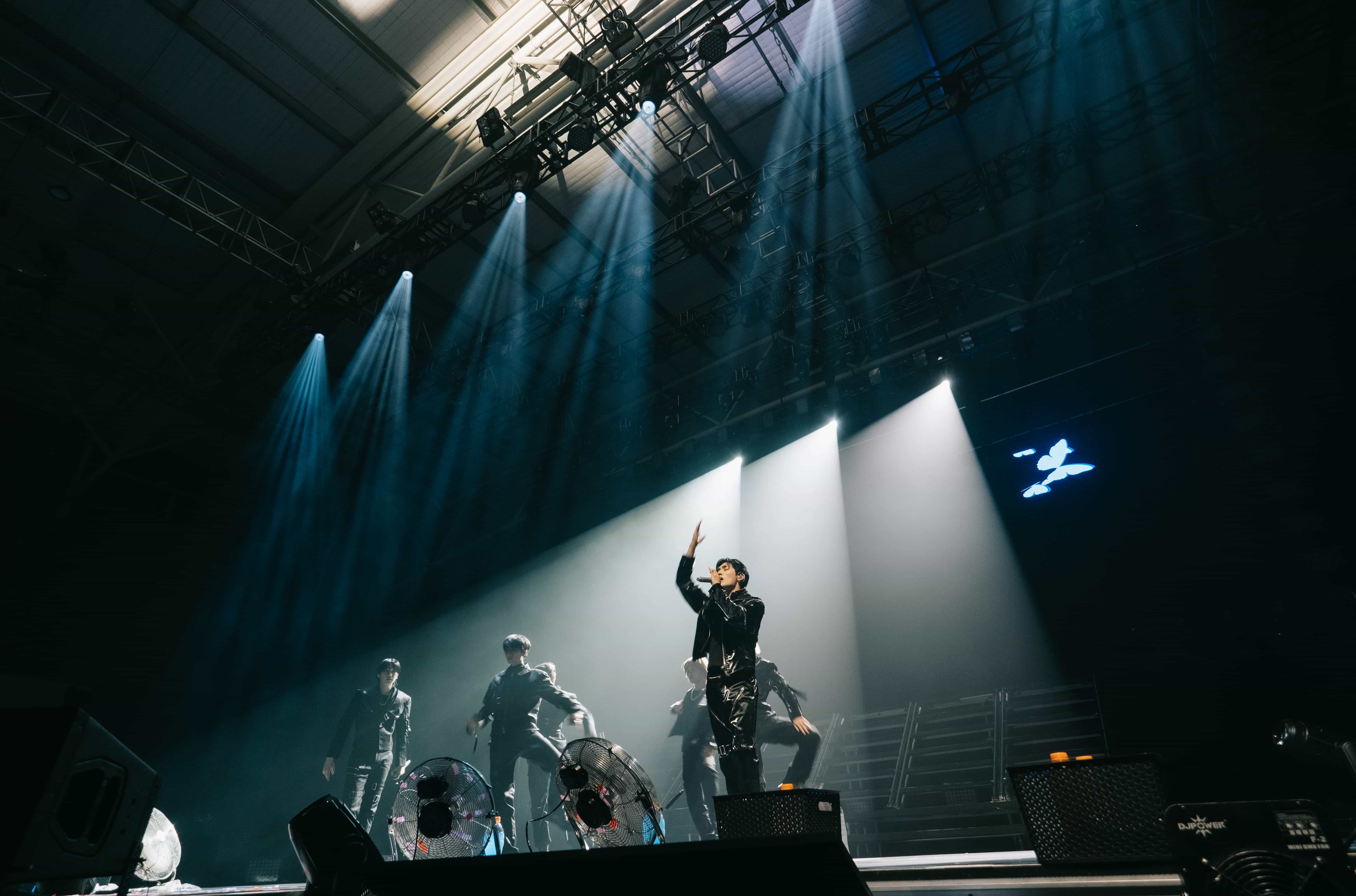 P1Harmony member pointing up from a raised platform as blue light beams sweep the arena