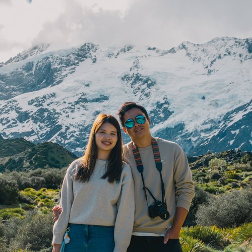 Julio and Kanis smiling side by side among alpine shrubs with snowy peaks behind
