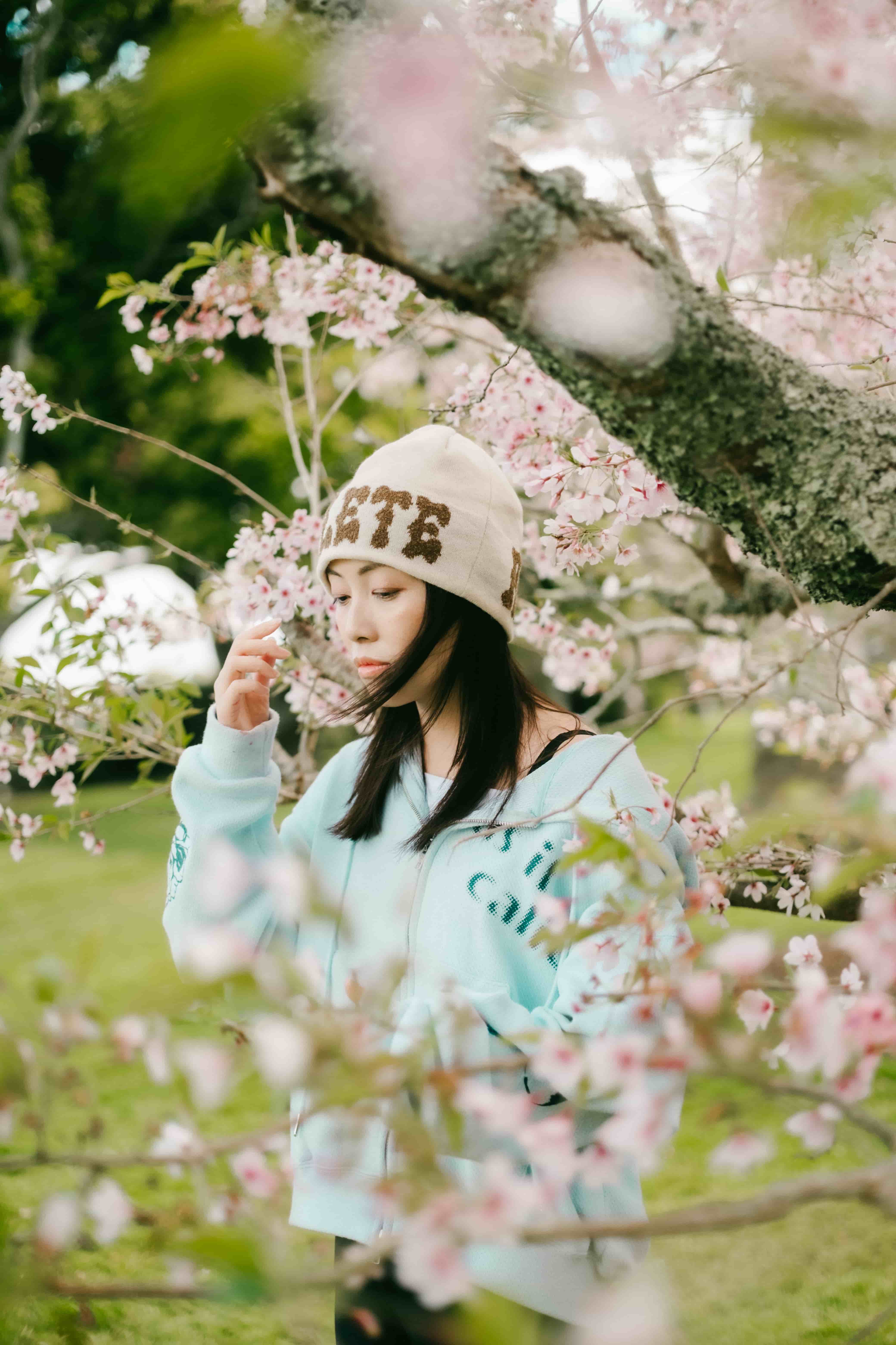 Young woman in a cream beanie and mint hoodie holding a cherry blossom twig up to her face