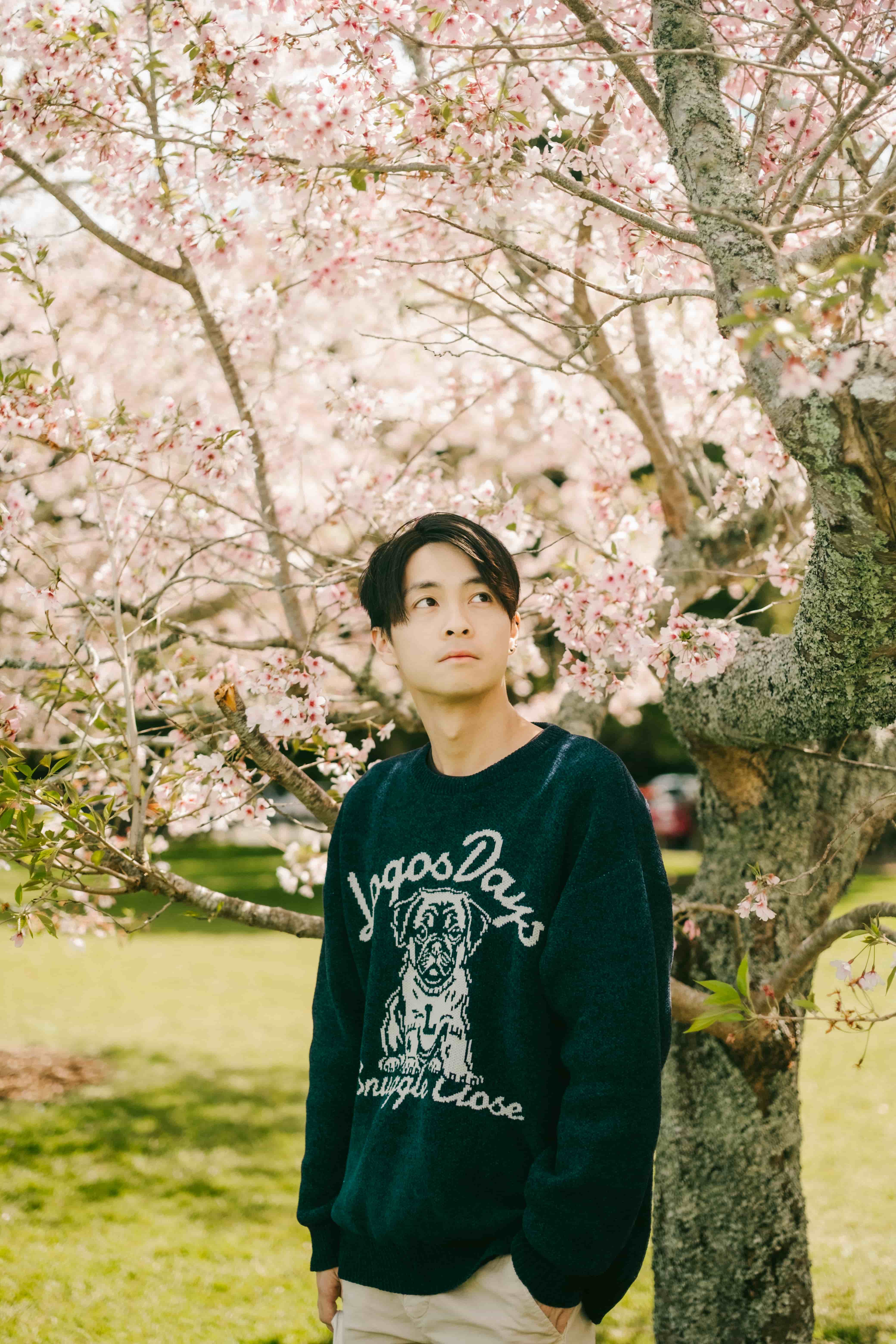 Young man in a navy sweater with his hands in his pockets beside a mossy cherry blossom tree