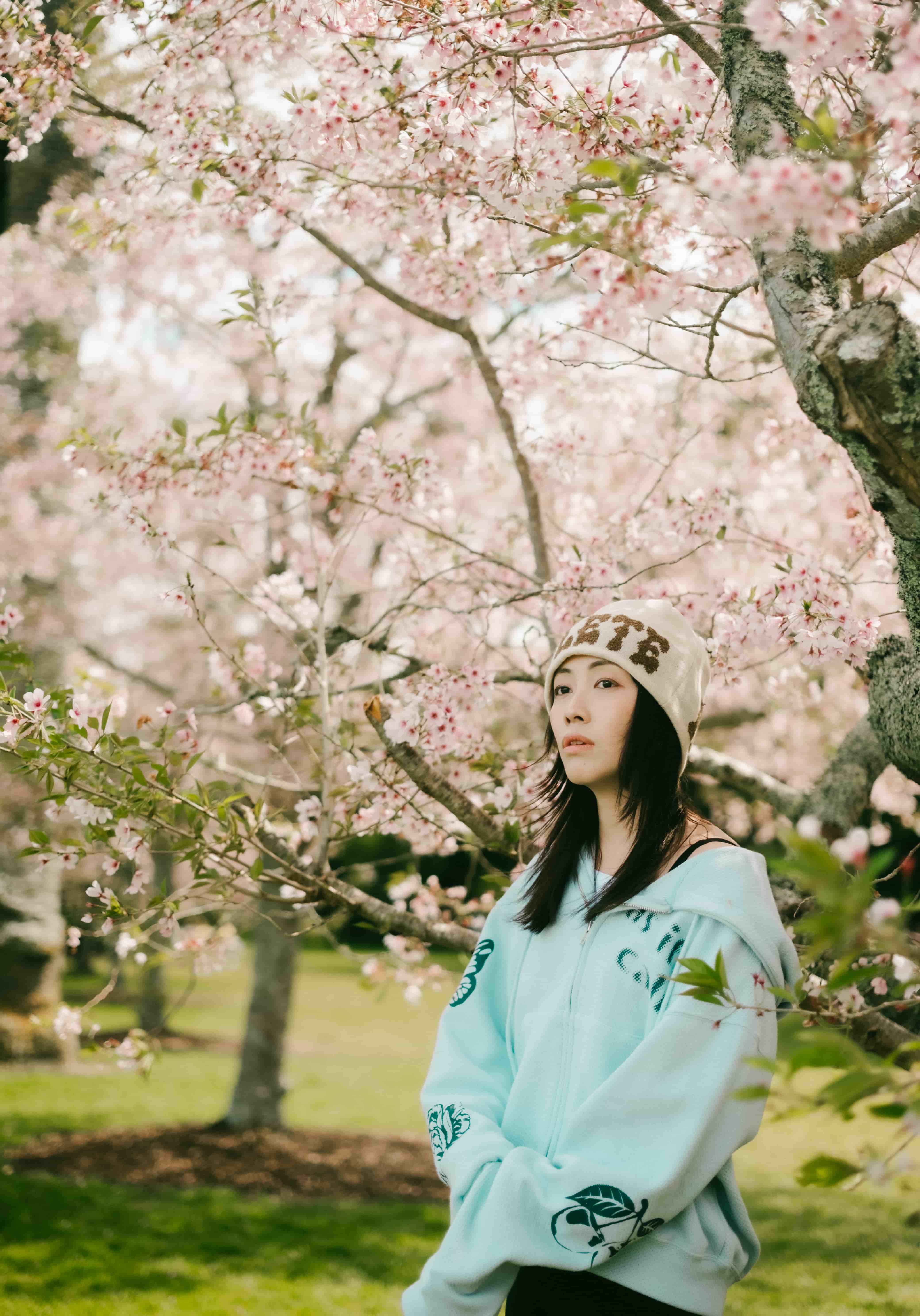 Young woman in a mint zip hoodie and beanie standing under a canopy of pink cherry blossom