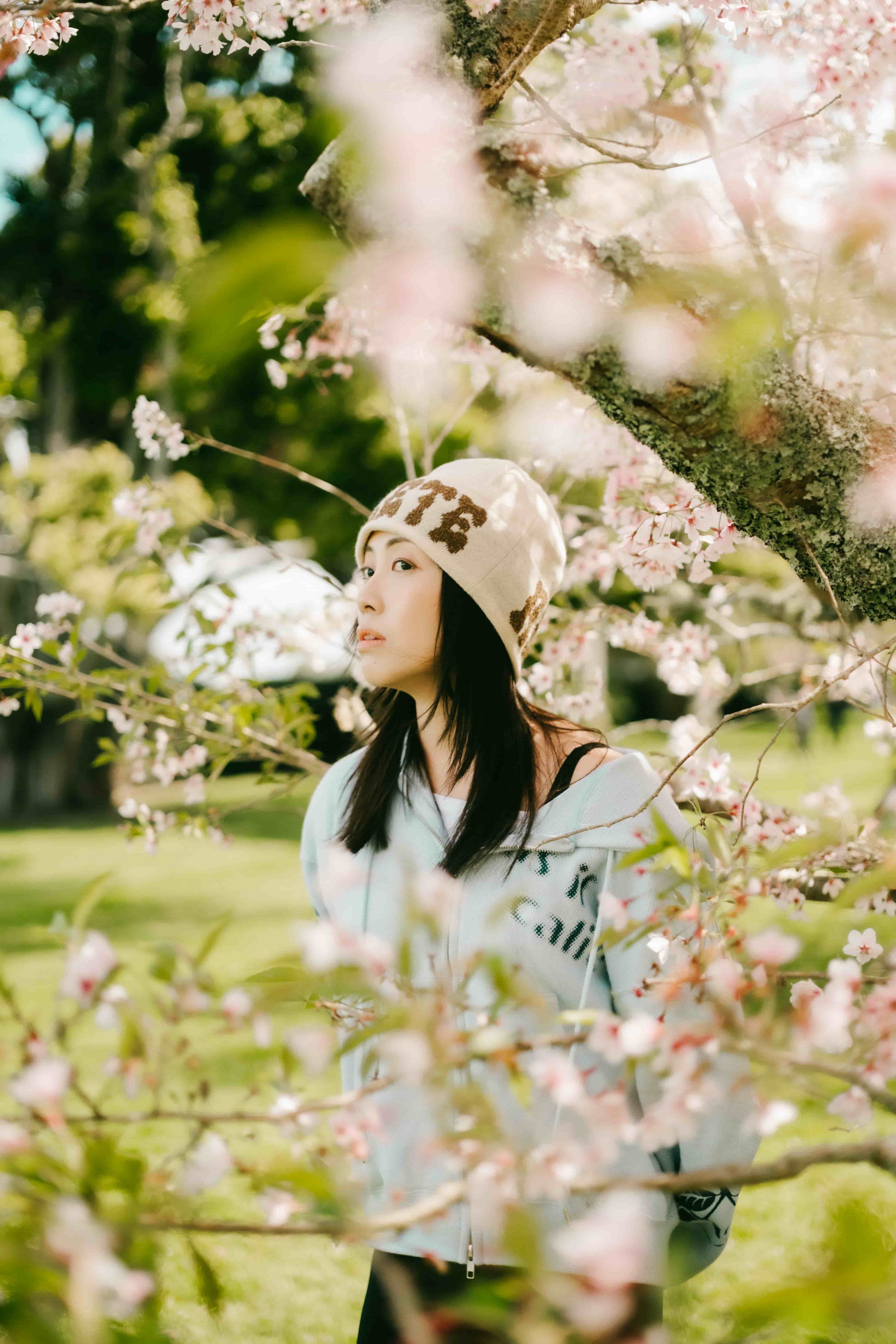 Young woman in a cream beanie looking up through soft, blurred pink cherry blossom