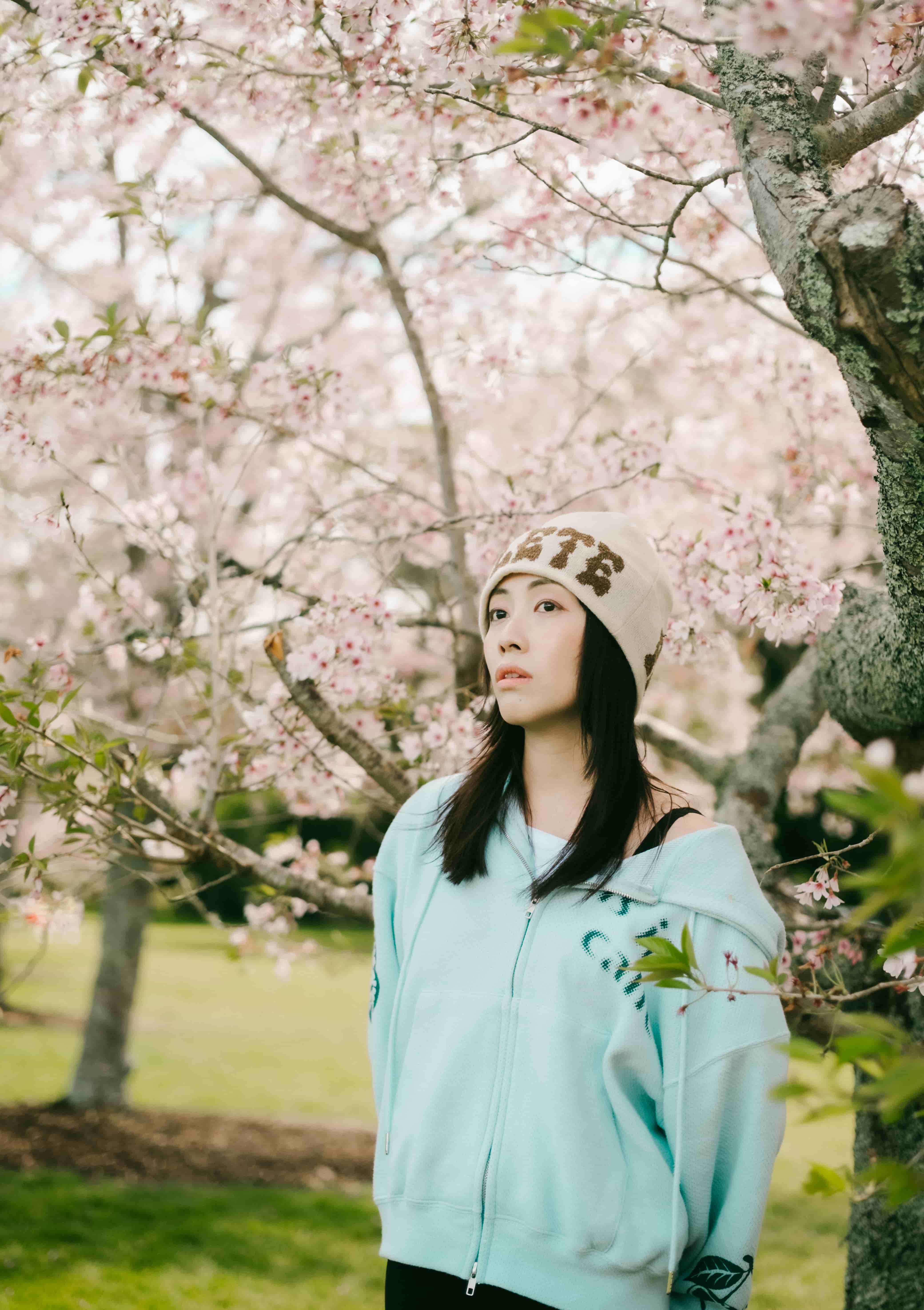 Young woman in a mint hoodie gazing upward beneath a large cherry blossom tree