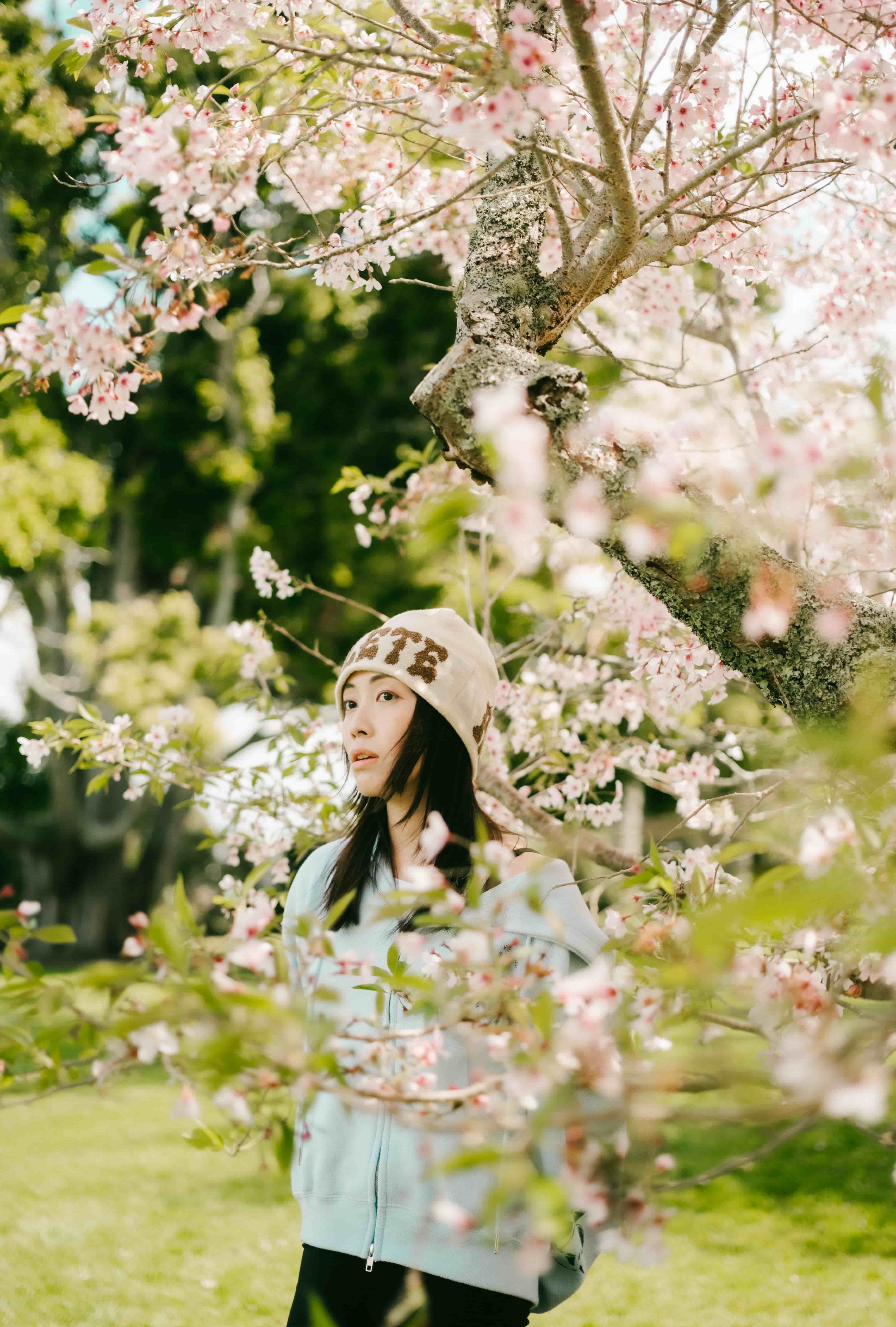 Young woman in a beanie and mint hoodie framed by pink blossom and green leaves