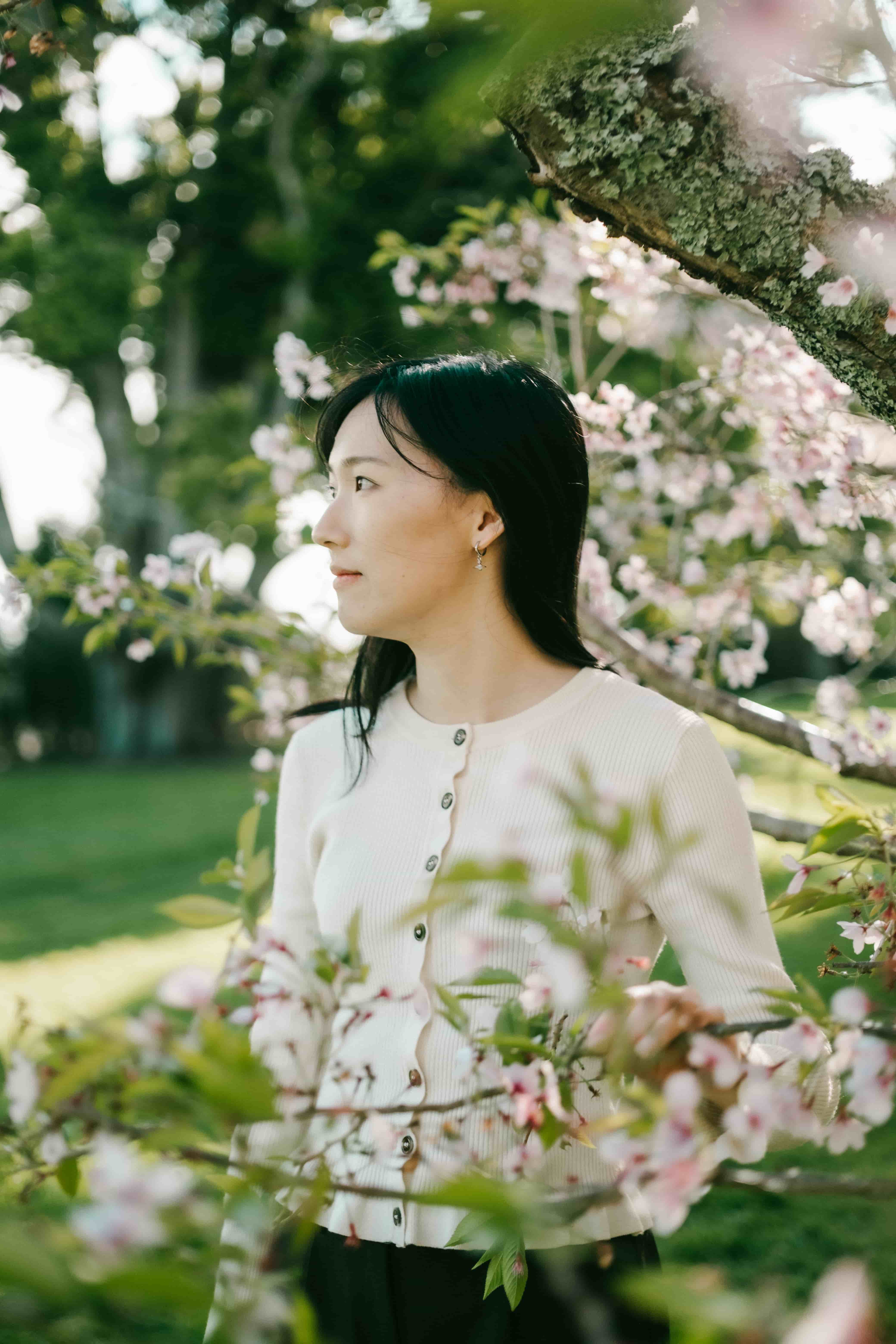 Young woman in a cream cardigan looking to the side, surrounded by cherry blossom and green leaves