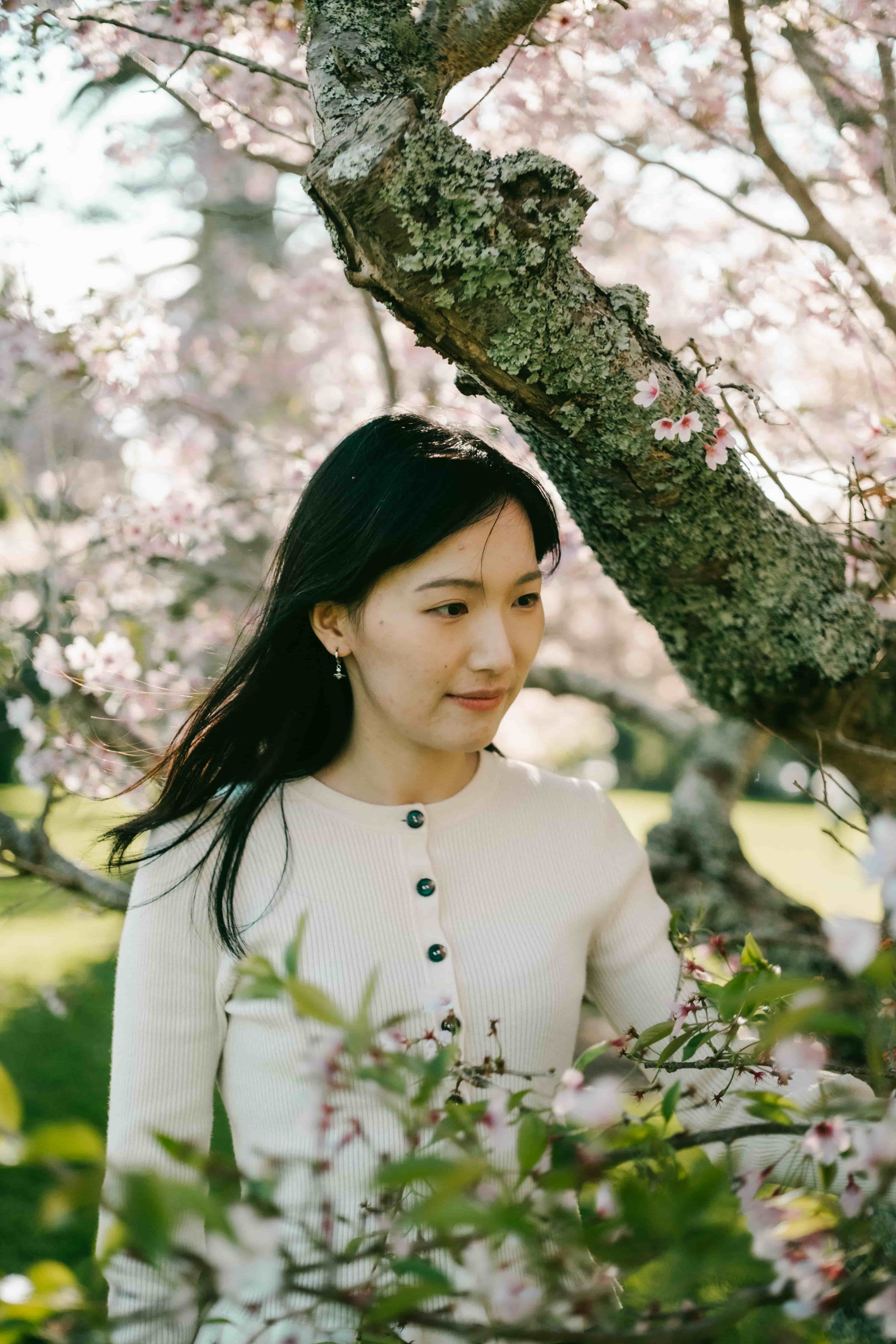 Young woman with long dark hair in a cream cardigan smiling softly under a mossy cherry blossom branch