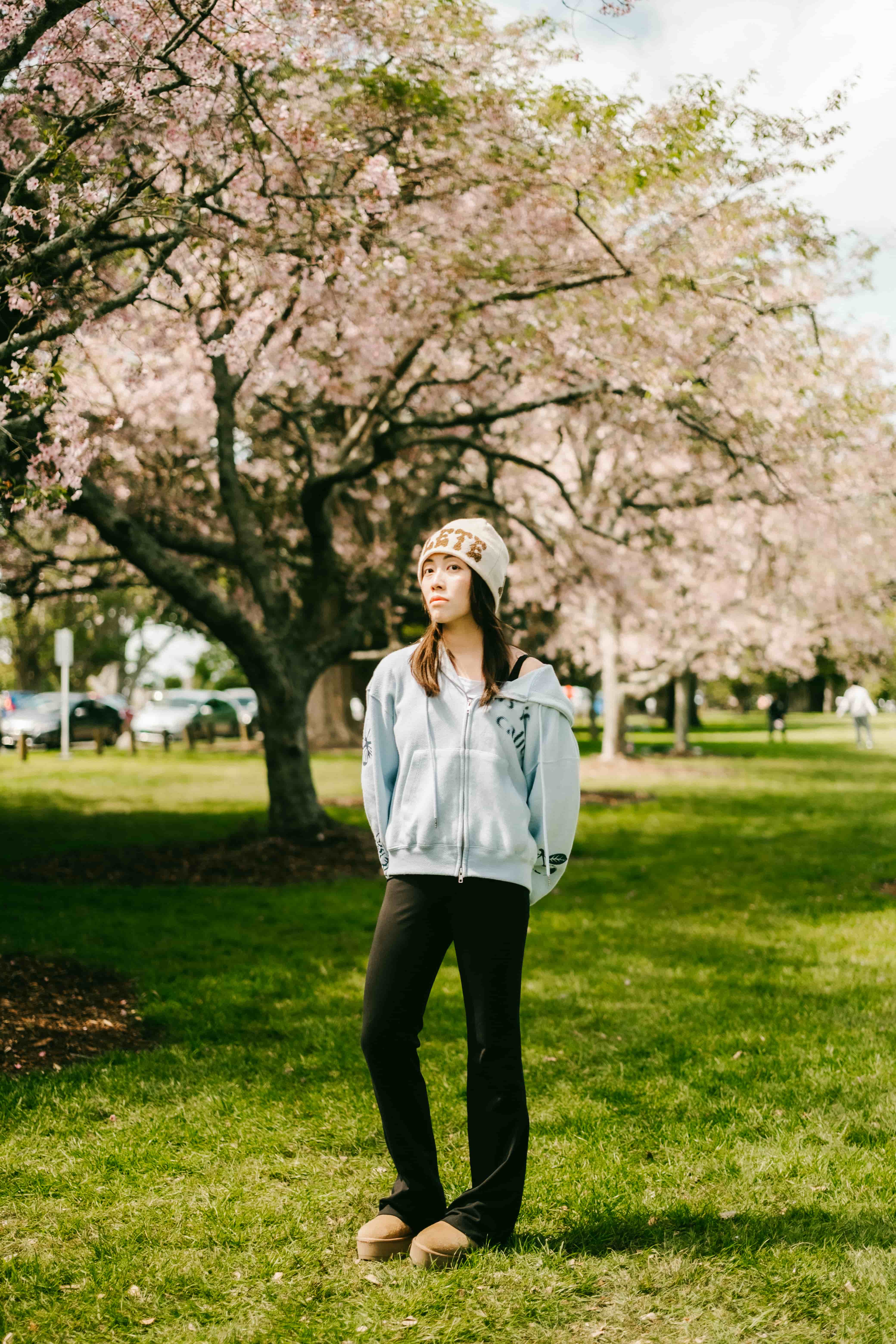 Young woman in a beanie, mint hoodie and black trousers standing on the lawn beneath cherry blossom trees