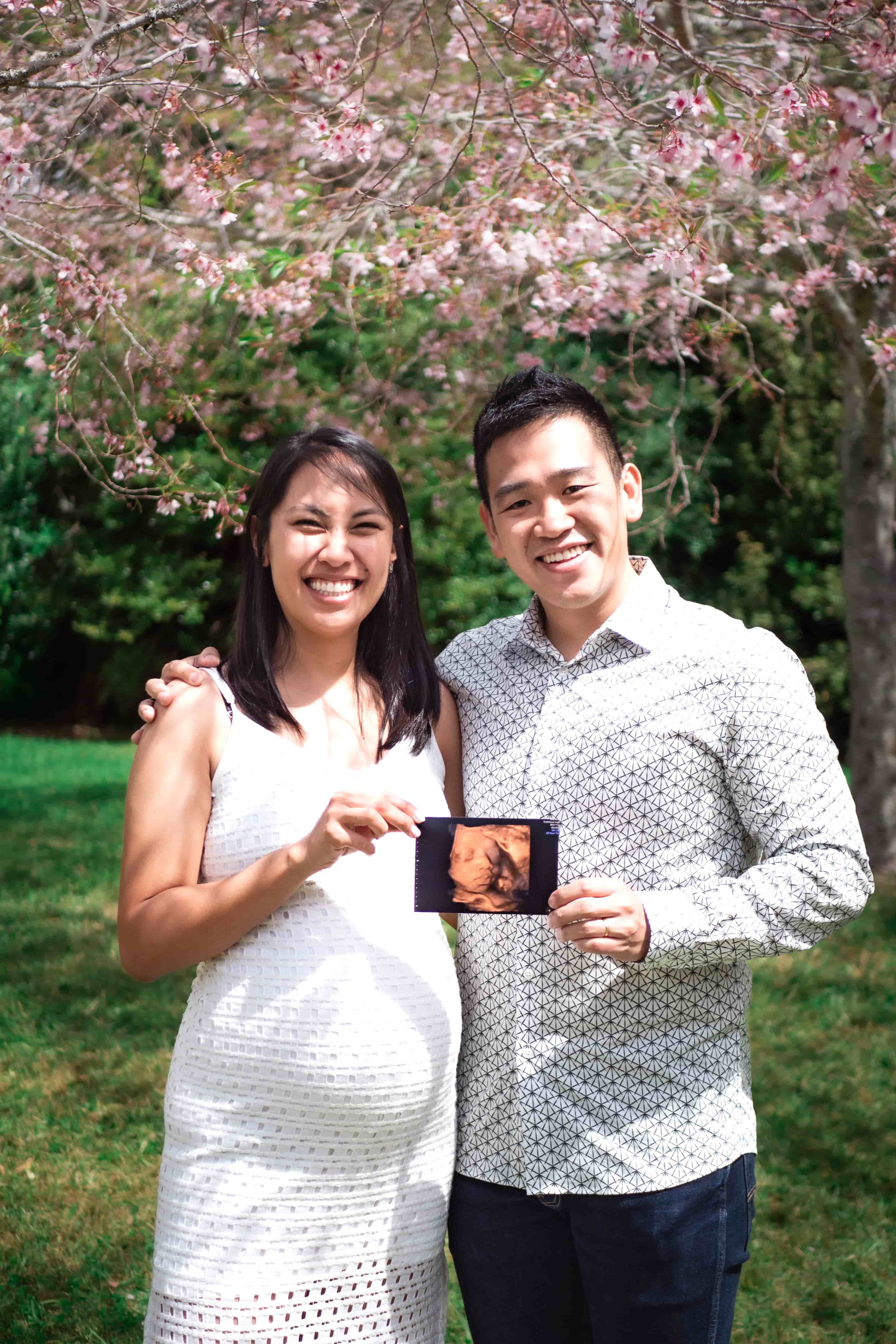 Lucas and Elisa holding up their baby scan photo under pink blossom
