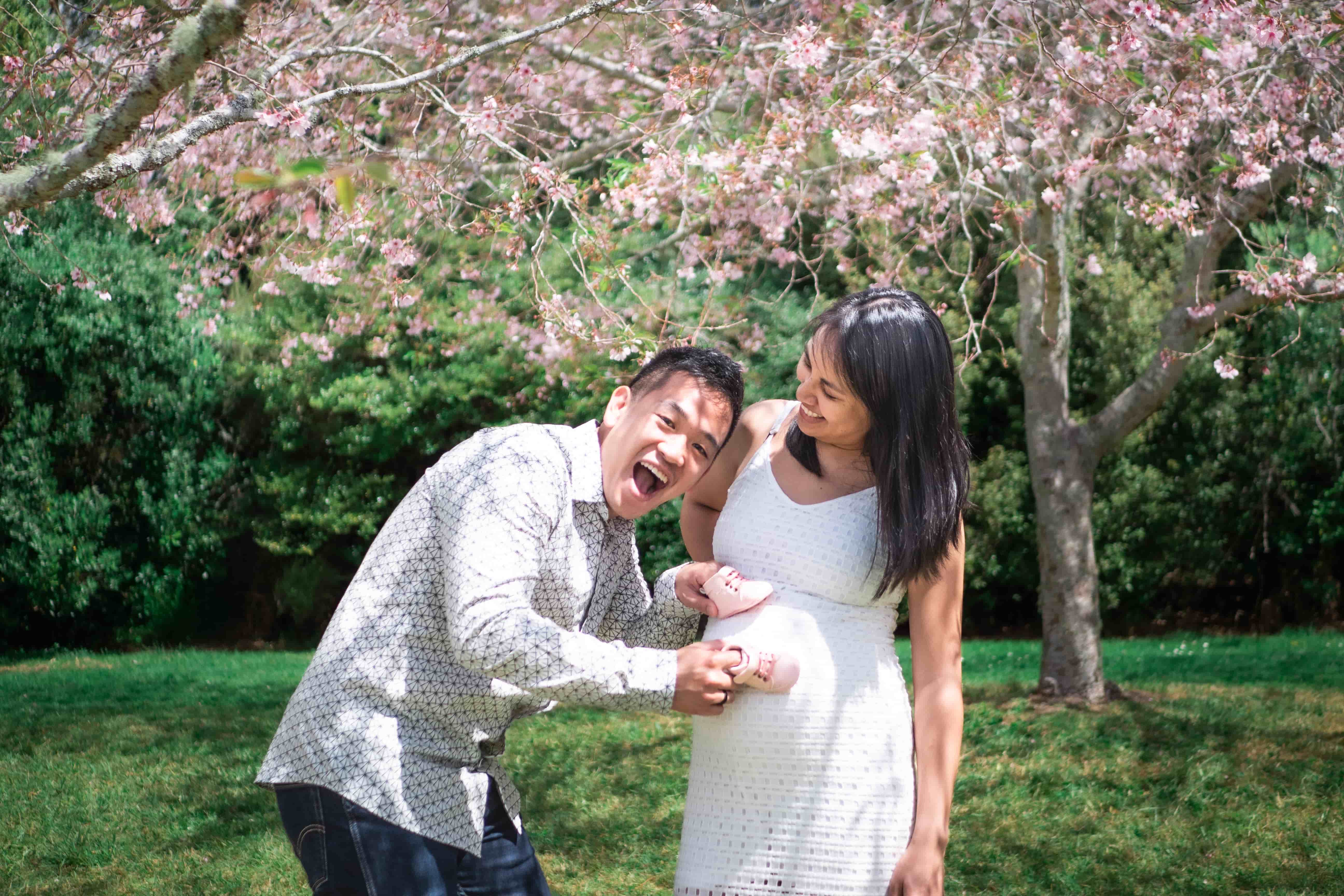 Lucas laughing as he holds the pink baby shoes to Elisa's bump under cherry blossom