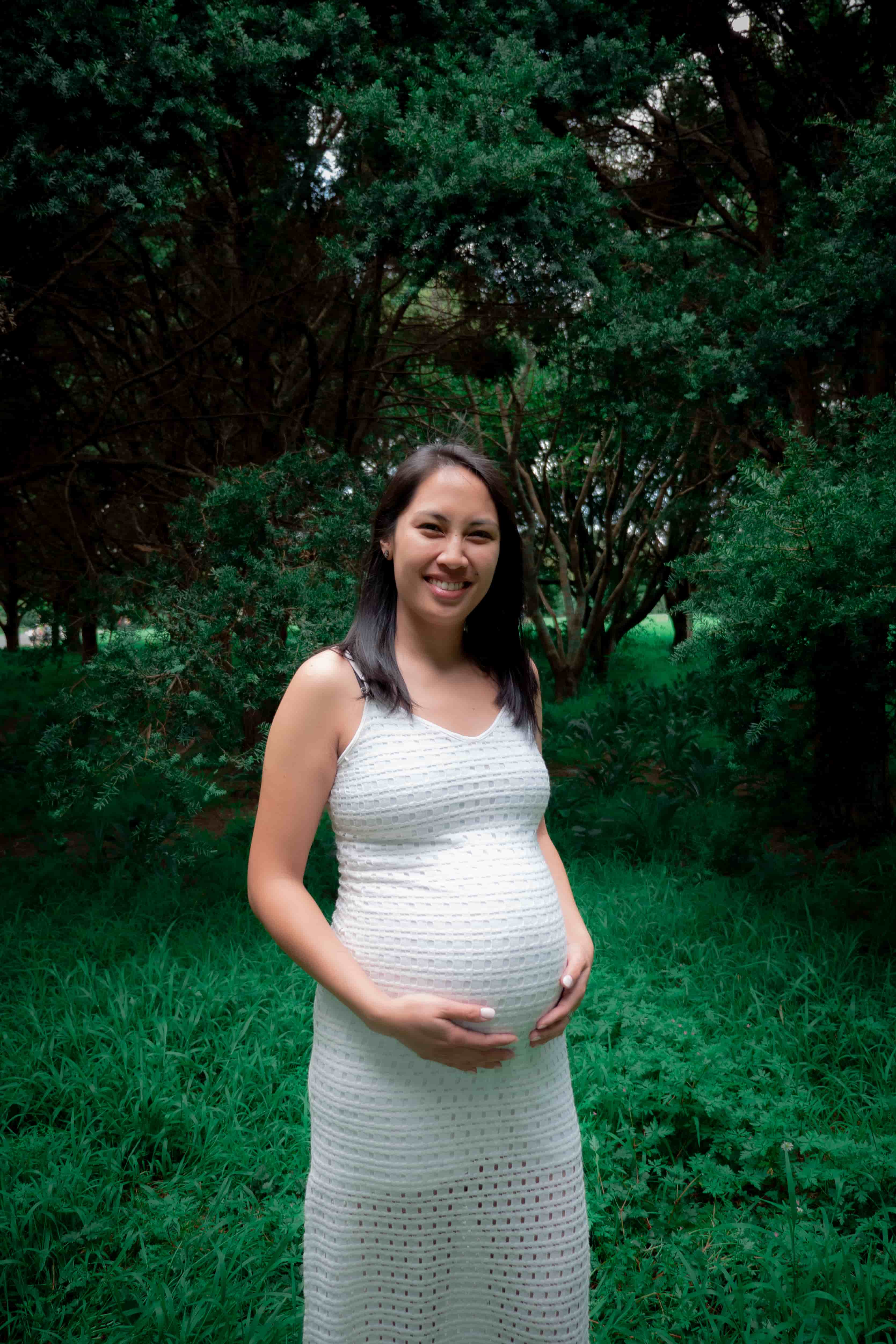 Elisa in a white dress cradling her bump among dark green trees