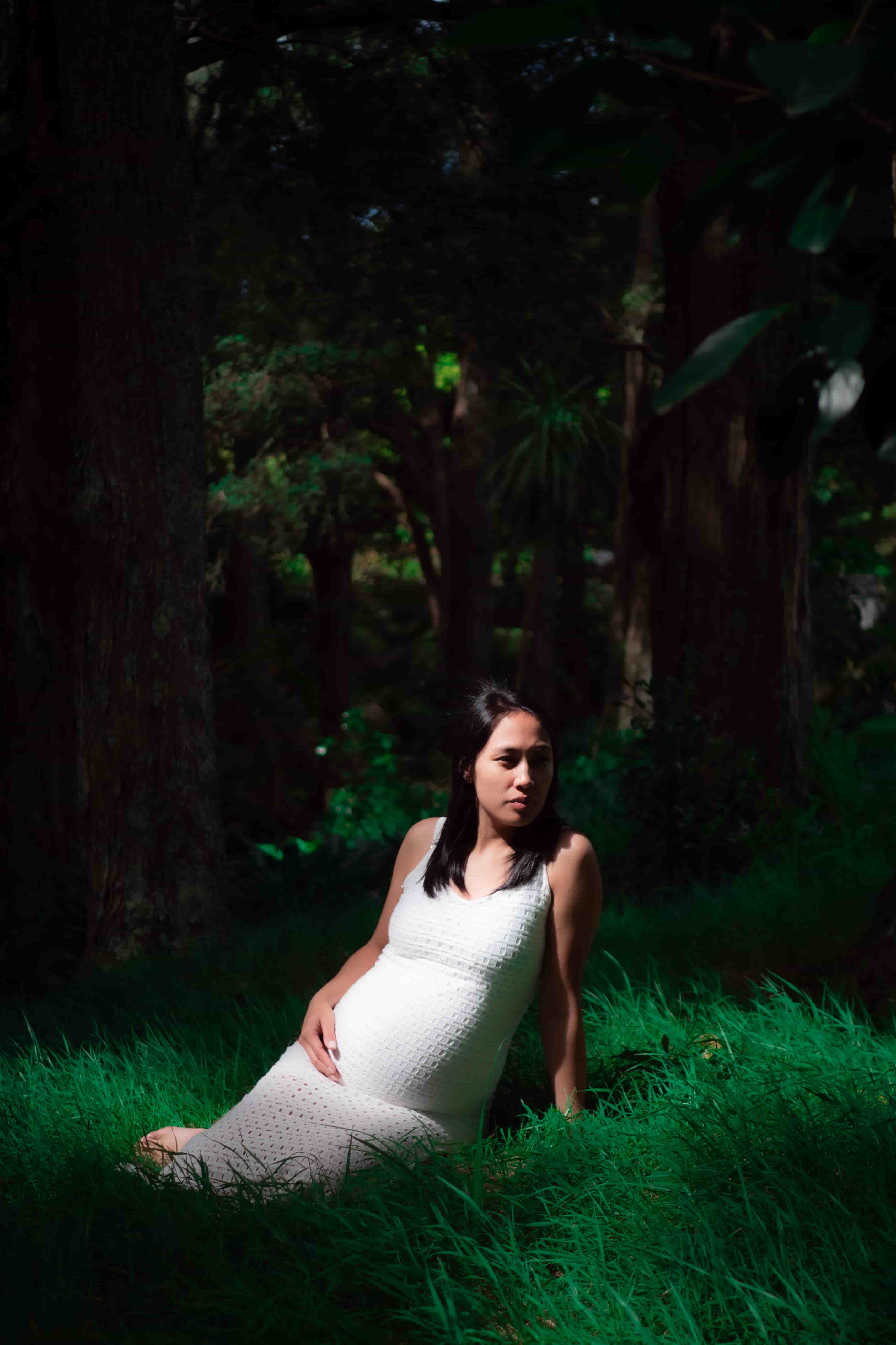 Elisa sitting in long grass in a white dress, shaded by tall trees