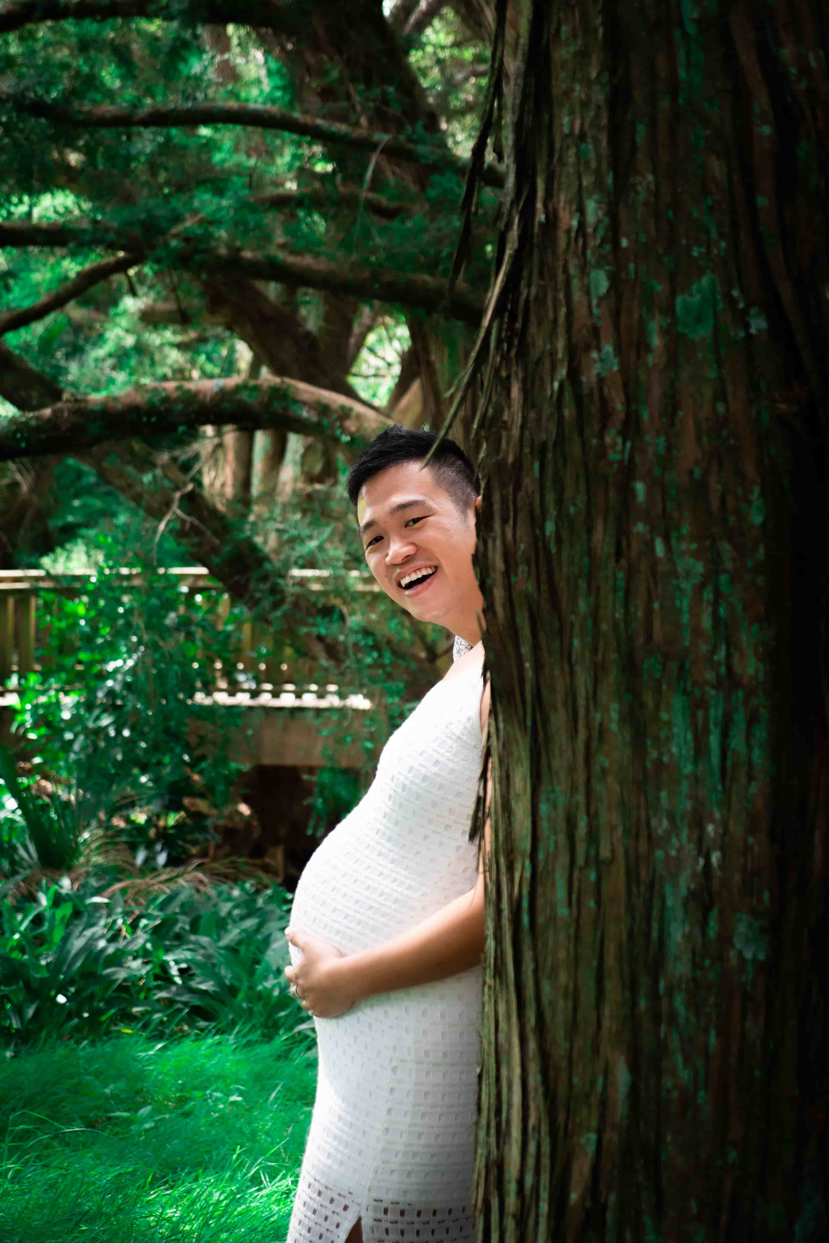 Playful shot of Lucas peeking out from behind a tree trunk above Elisa's bump, as if he is the one expecting