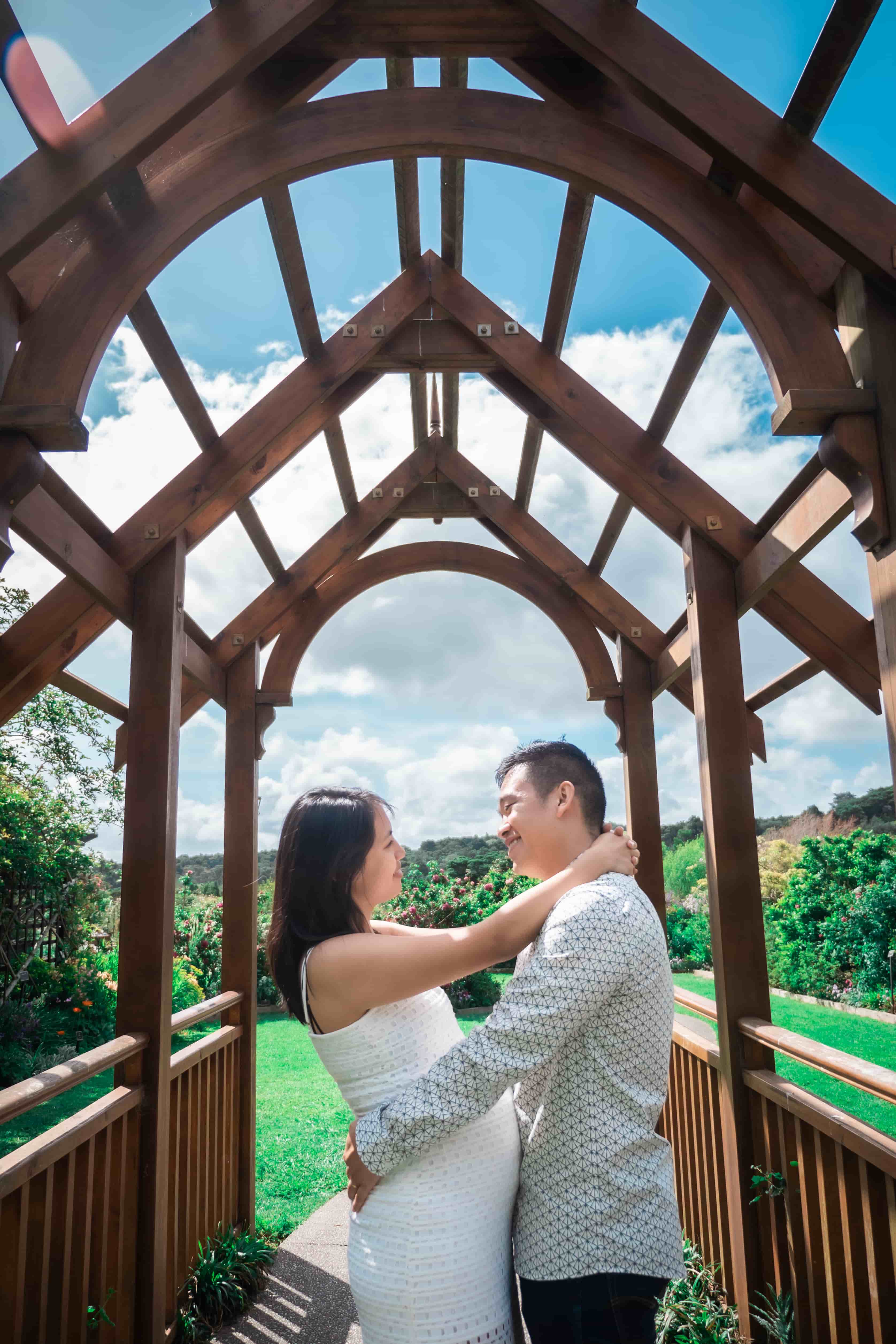 Lucas and Elisa embracing under a wooden arched walkway and blue sky