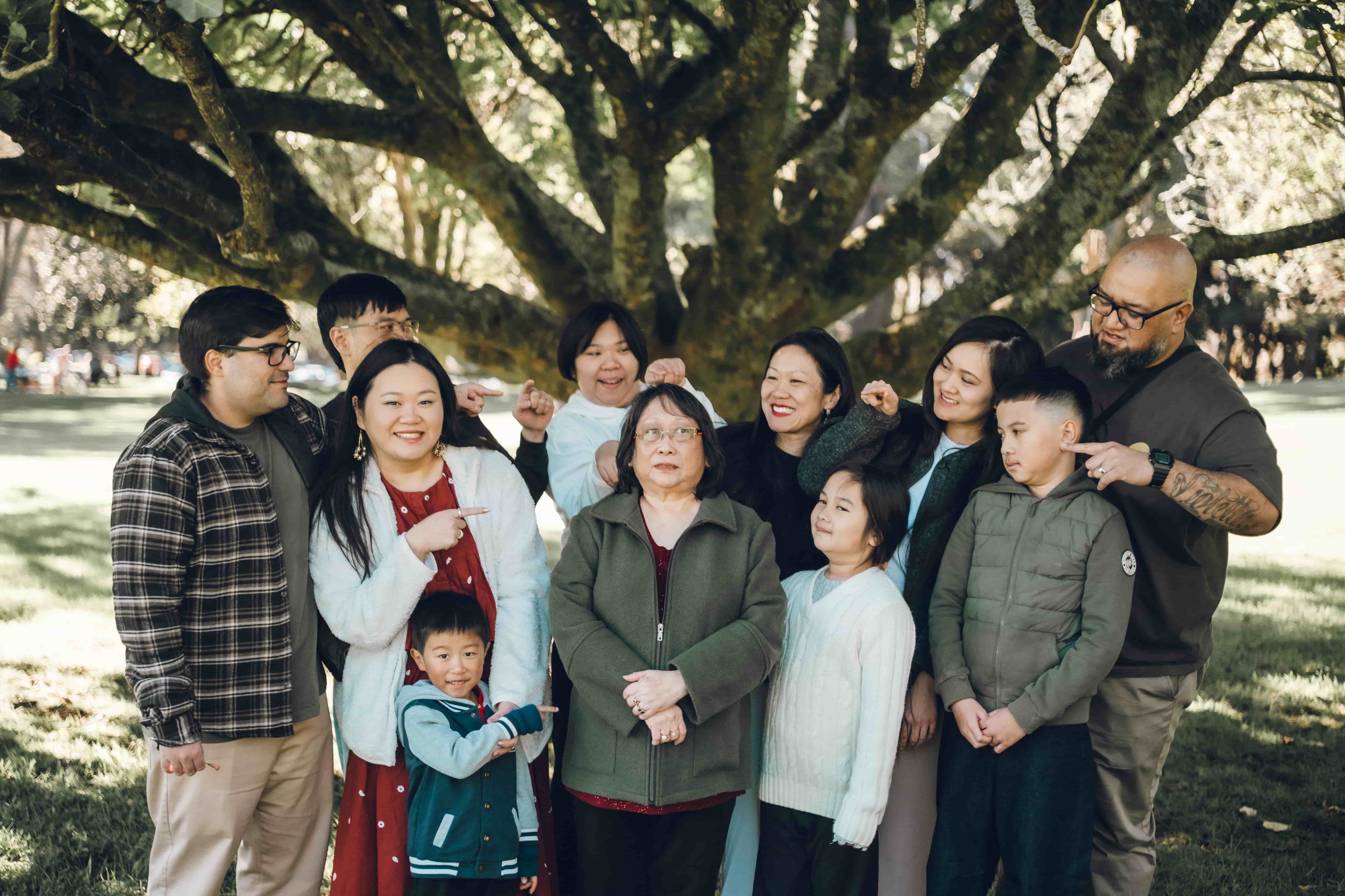 Extended family playfully pointing at grandma under a big tree, the youngest grandson looking up at her