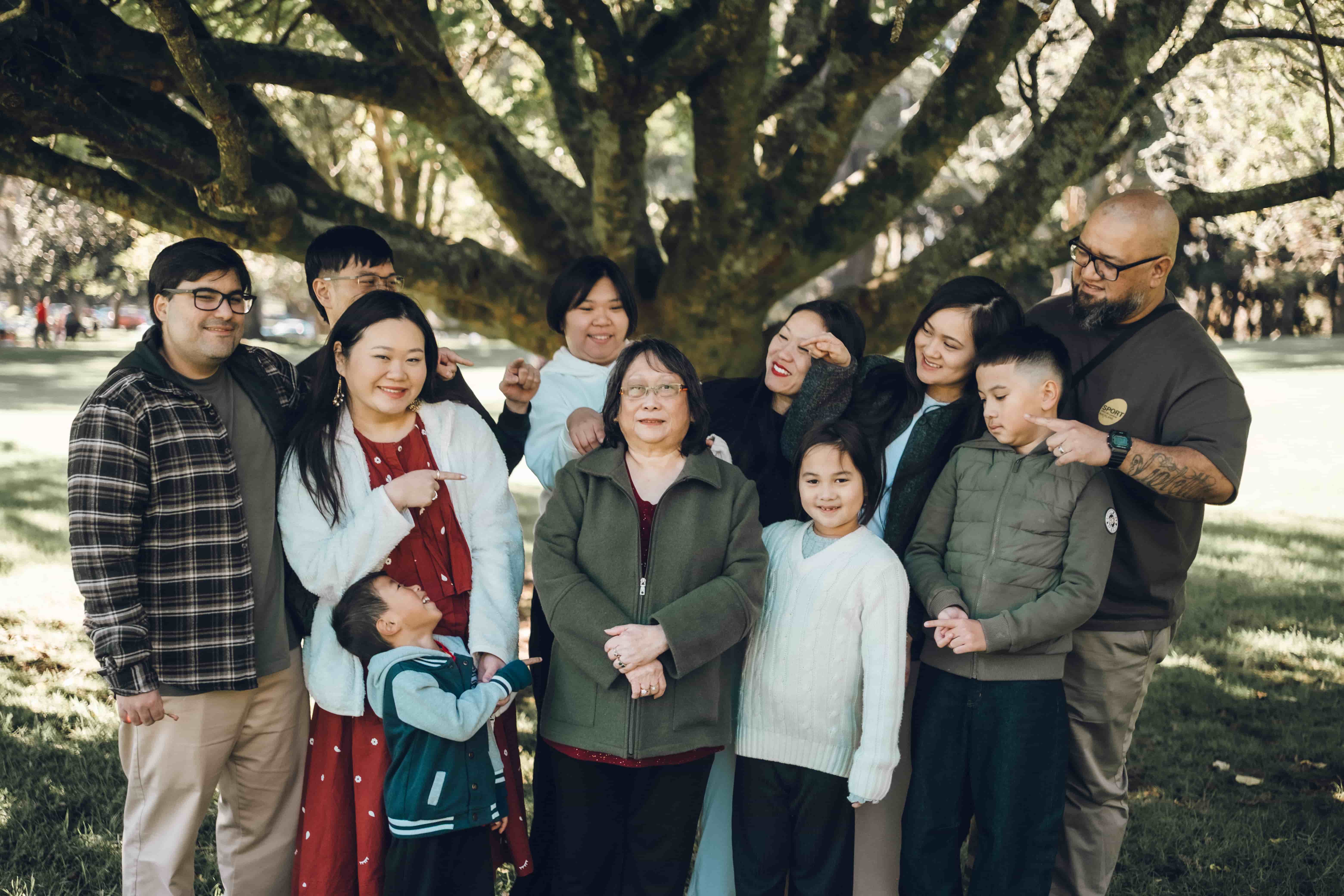 Extended family playfully pointing at grandma under a big tree, the youngest grandson looking up at her
