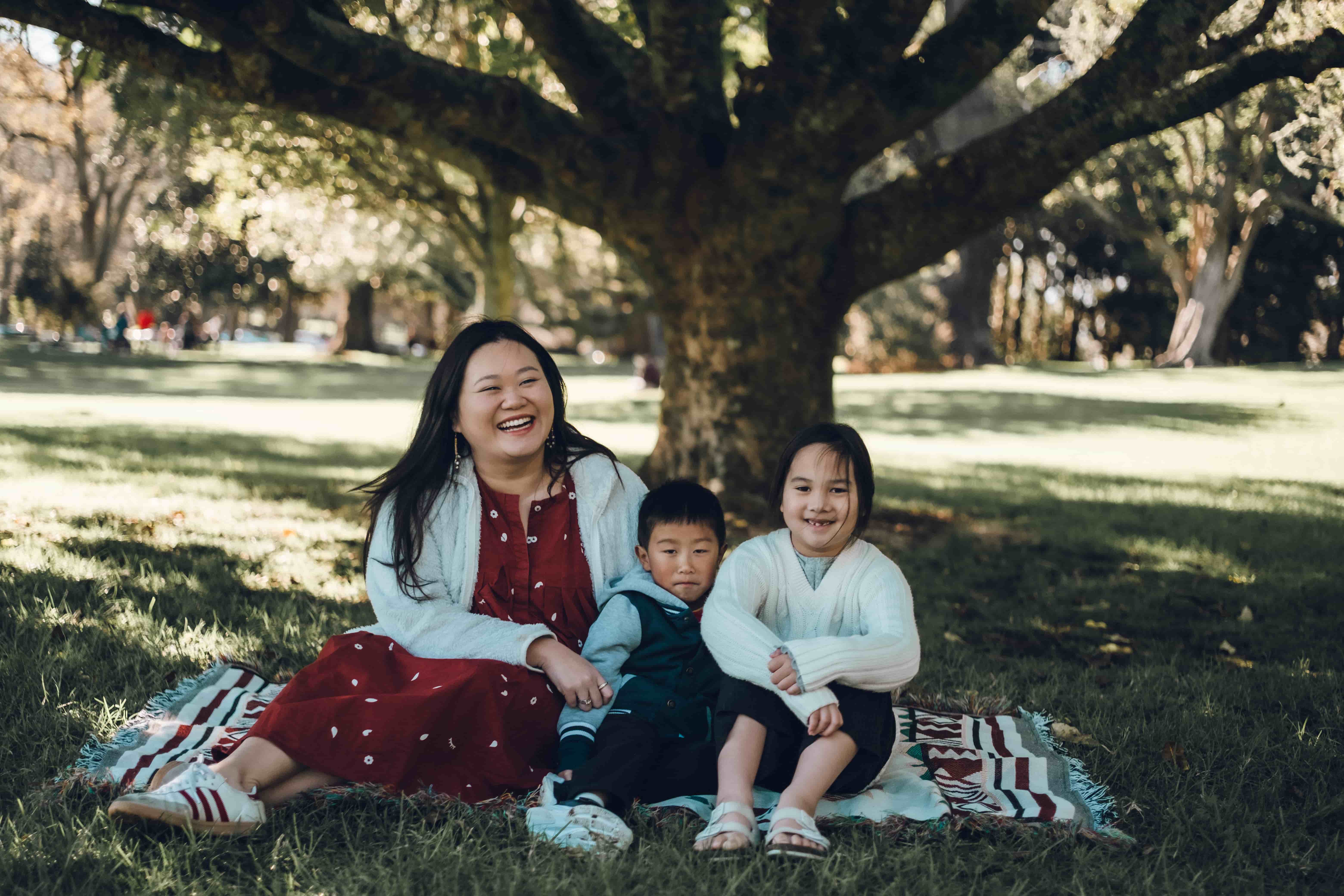 Mother hugging two children on a blanket beneath spreading branches