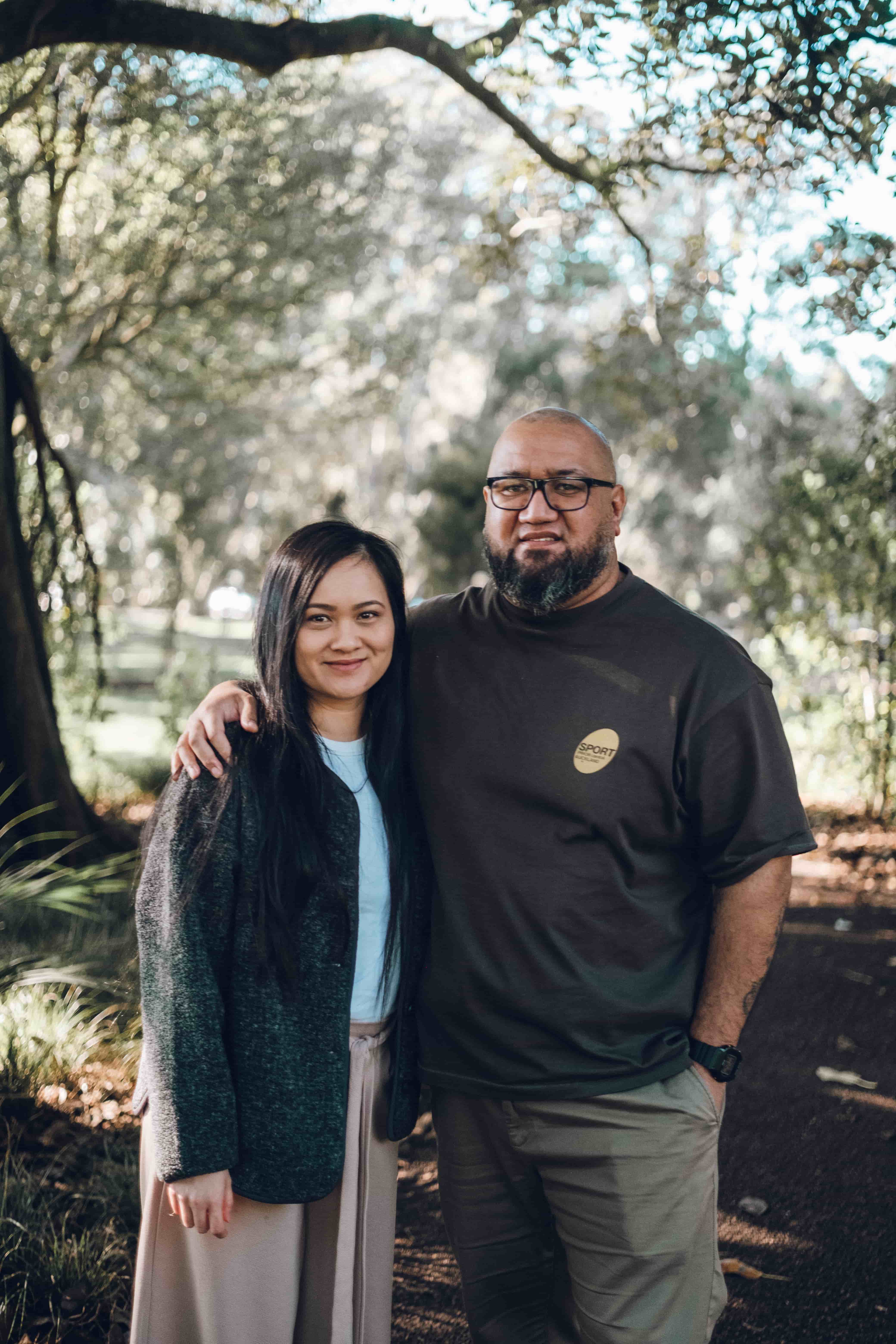 Couple smiling, her arm around his shoulder, trees behind them