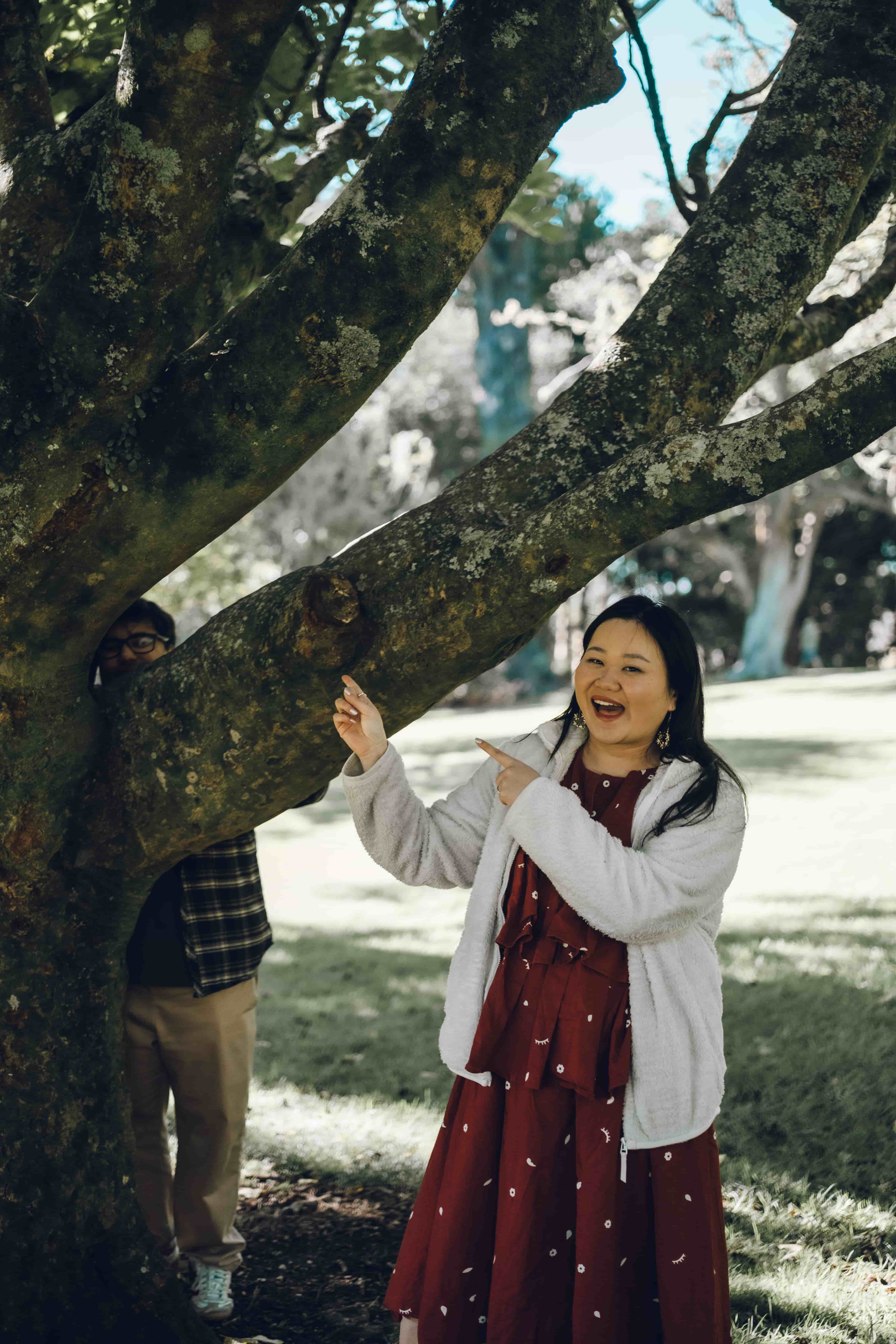 Woman laughing and pointing as her partner hides behind a tree trunk