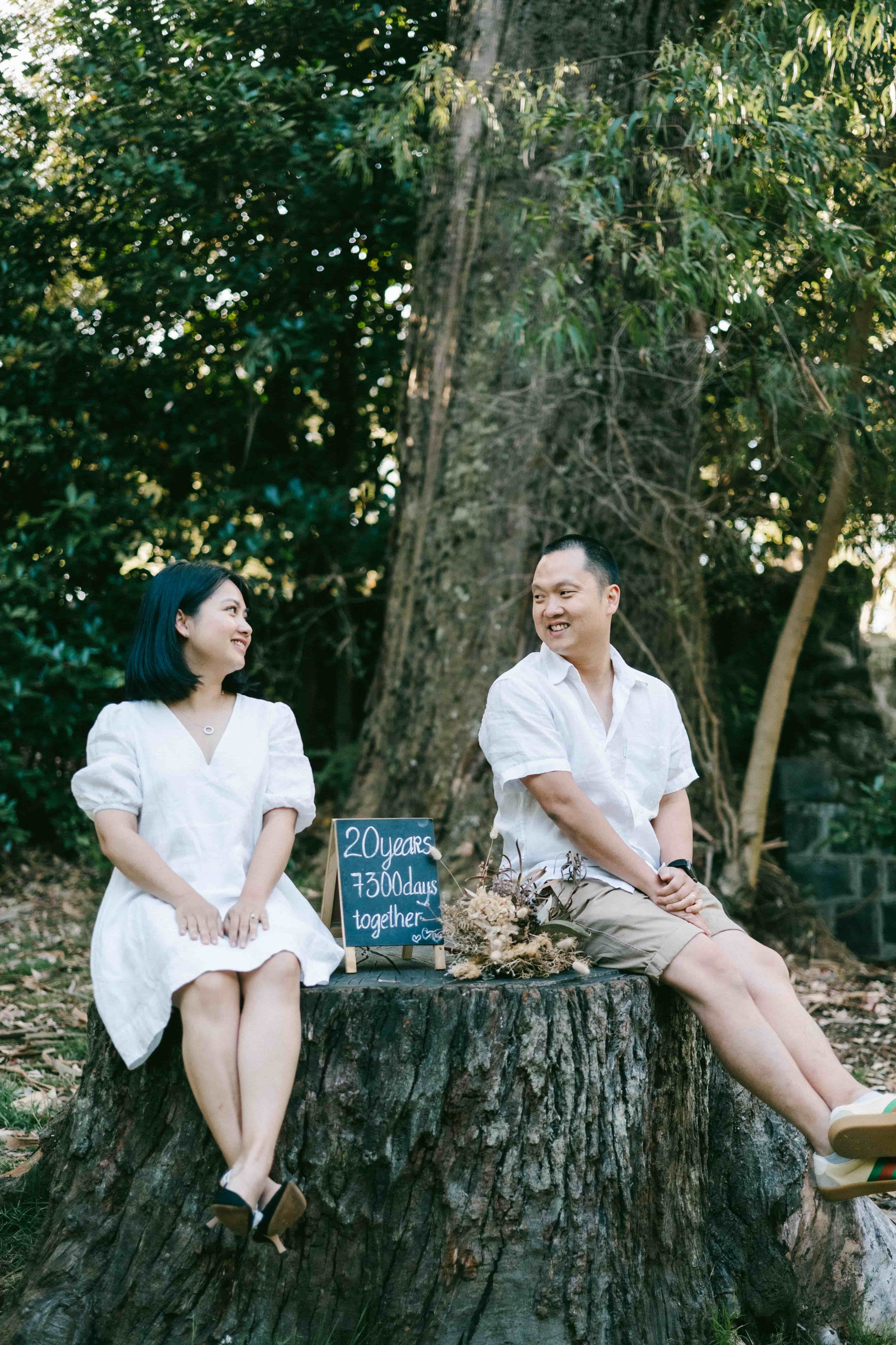 Couple sitting on a tree stump smiling at each other, the chalkboard between them