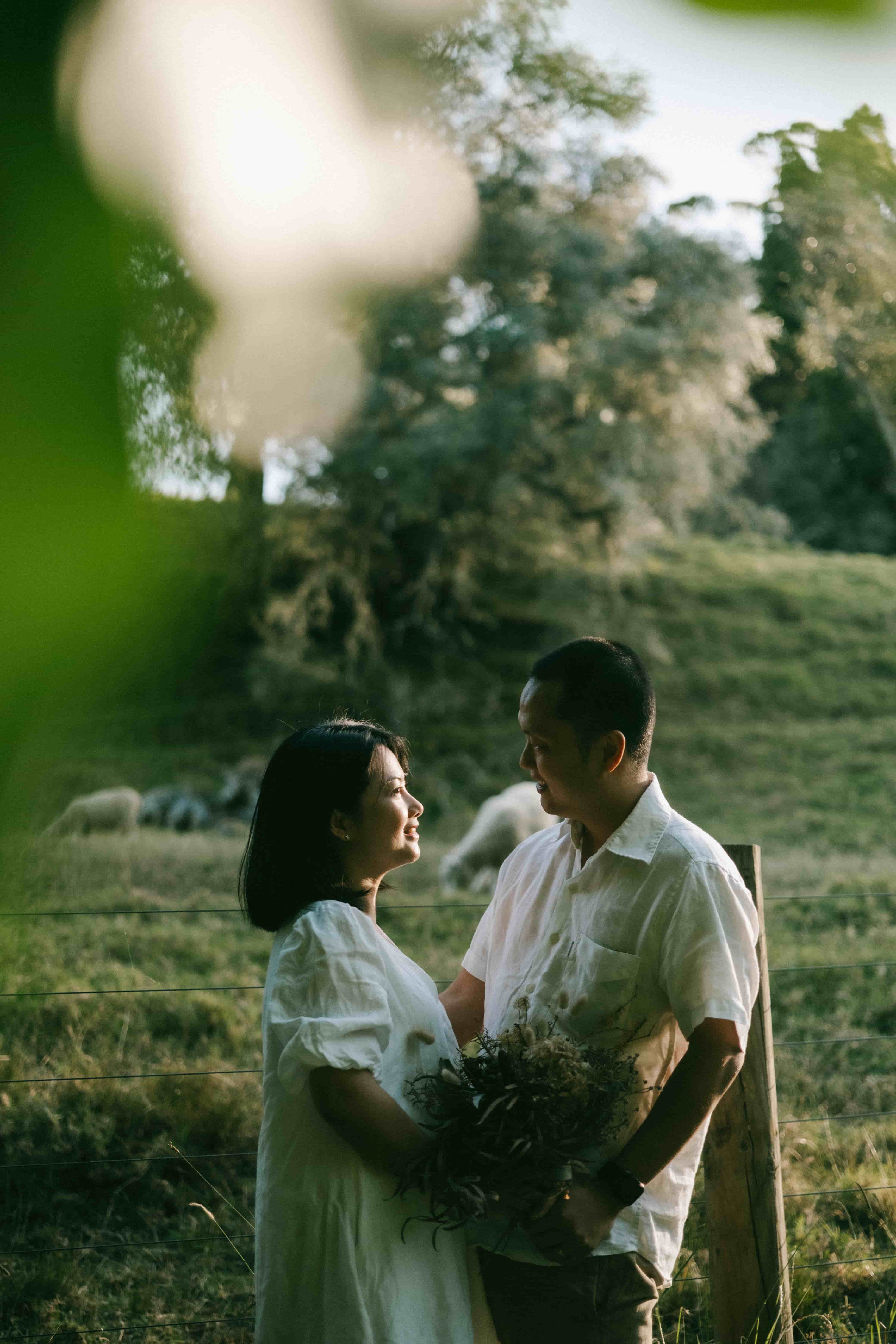 Couple facing each other by a fence, sheep grazing in the paddock behind