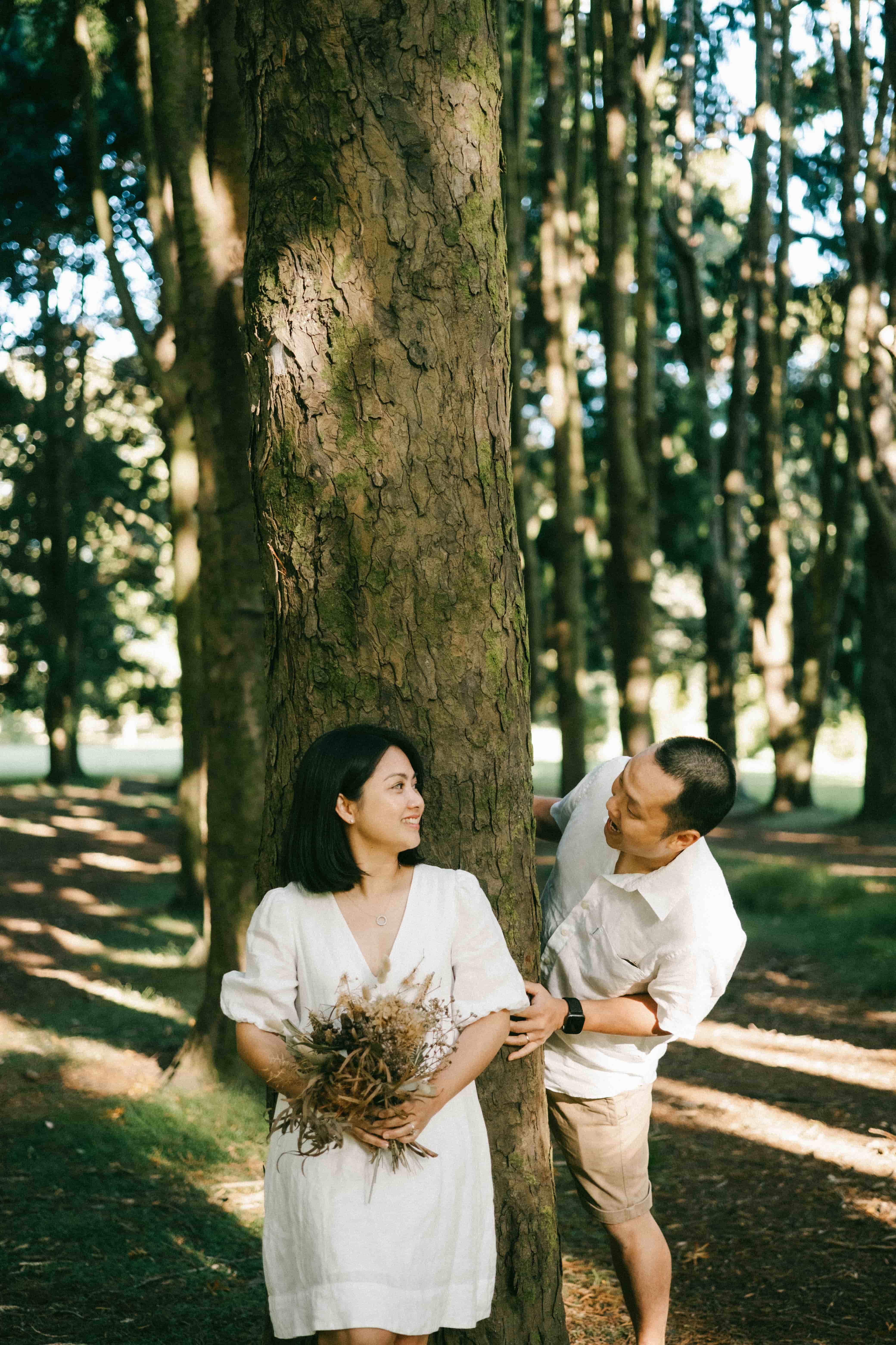 Wife holding the bouquet in front of a tree as her husband peeks out