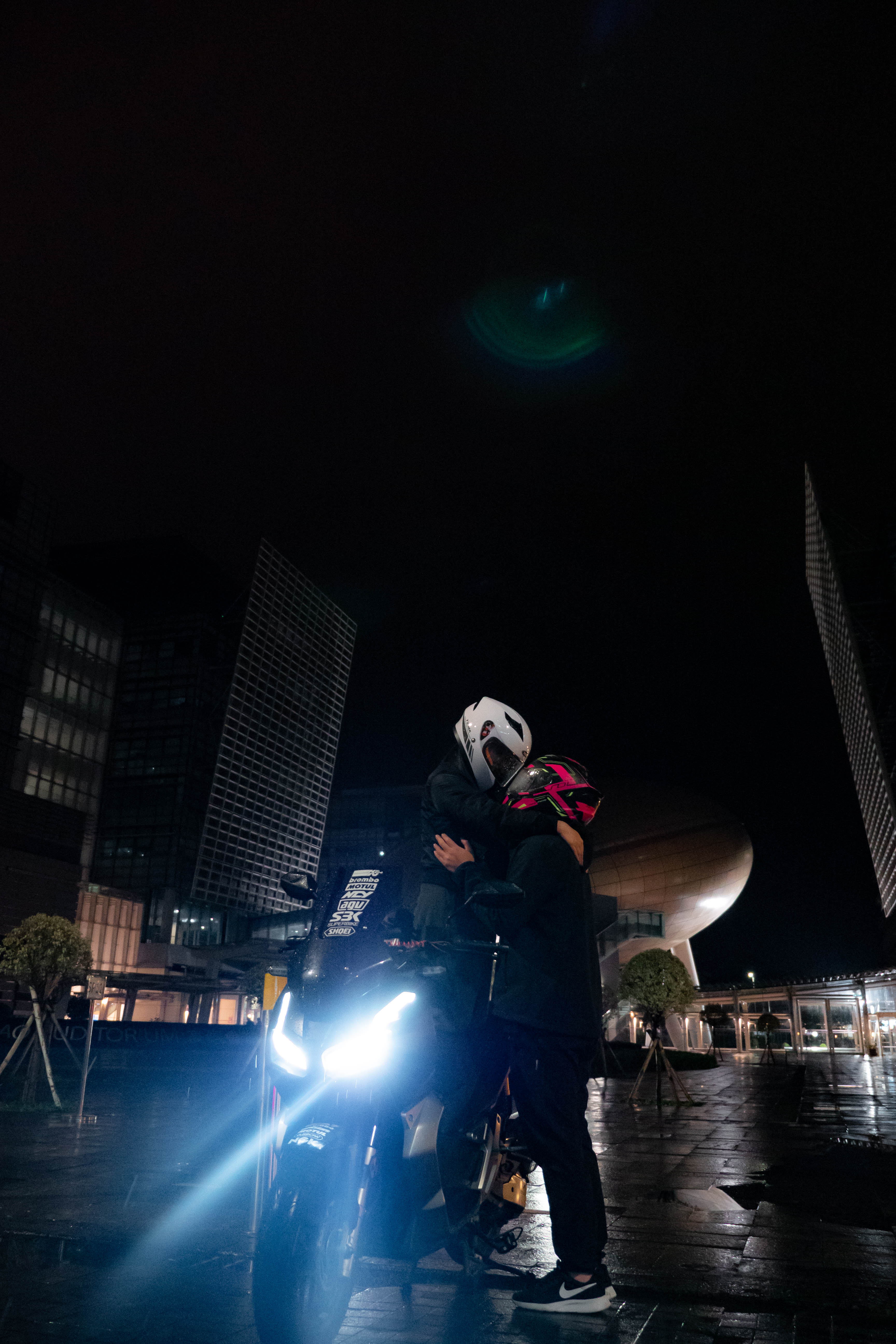Two riders in helmets embracing beside the glowing motorcycle, glass towers and a domed building behind