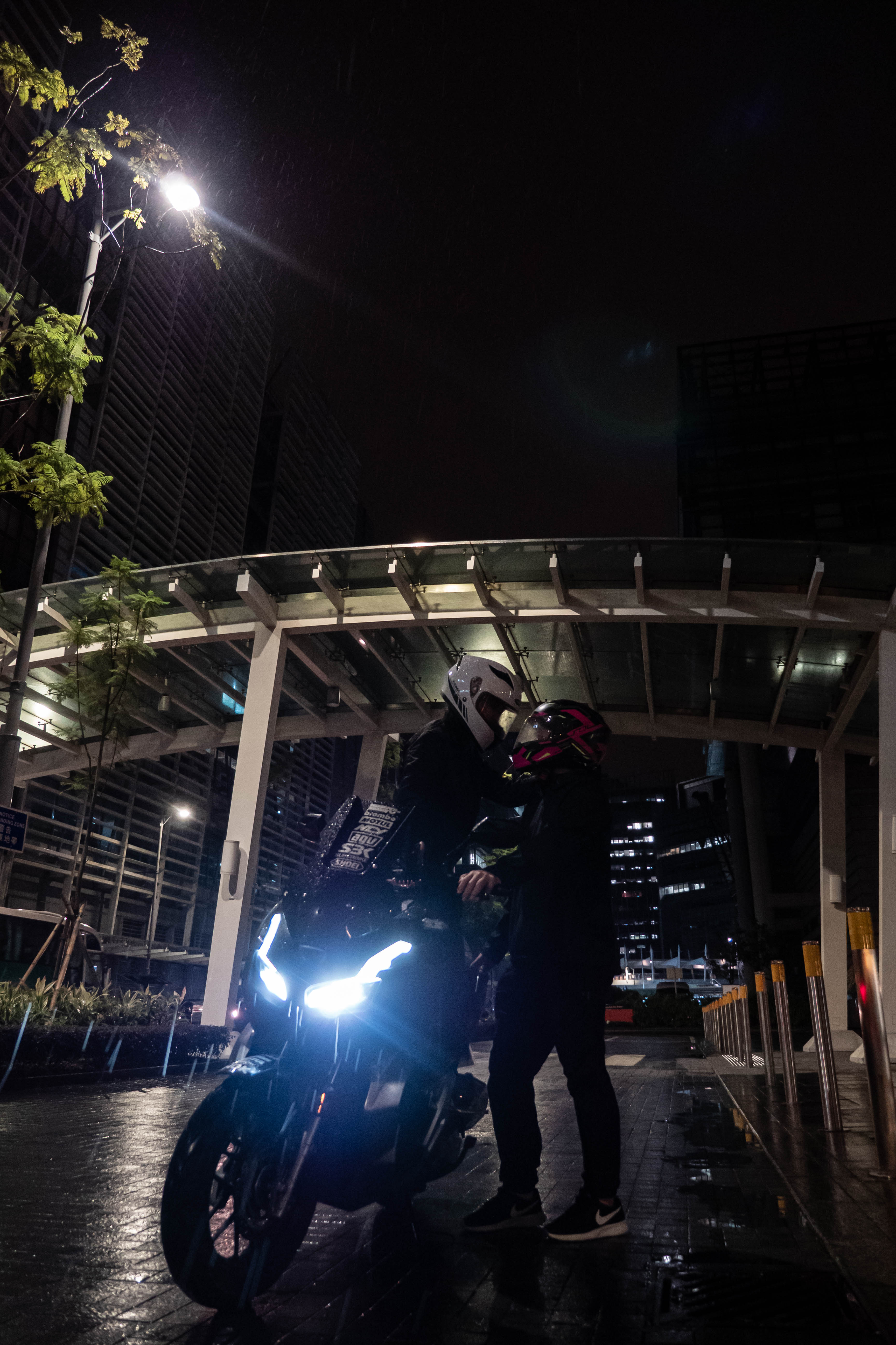 Rider standing beside the motorcycle at night, its headlight bright on a wet plaza