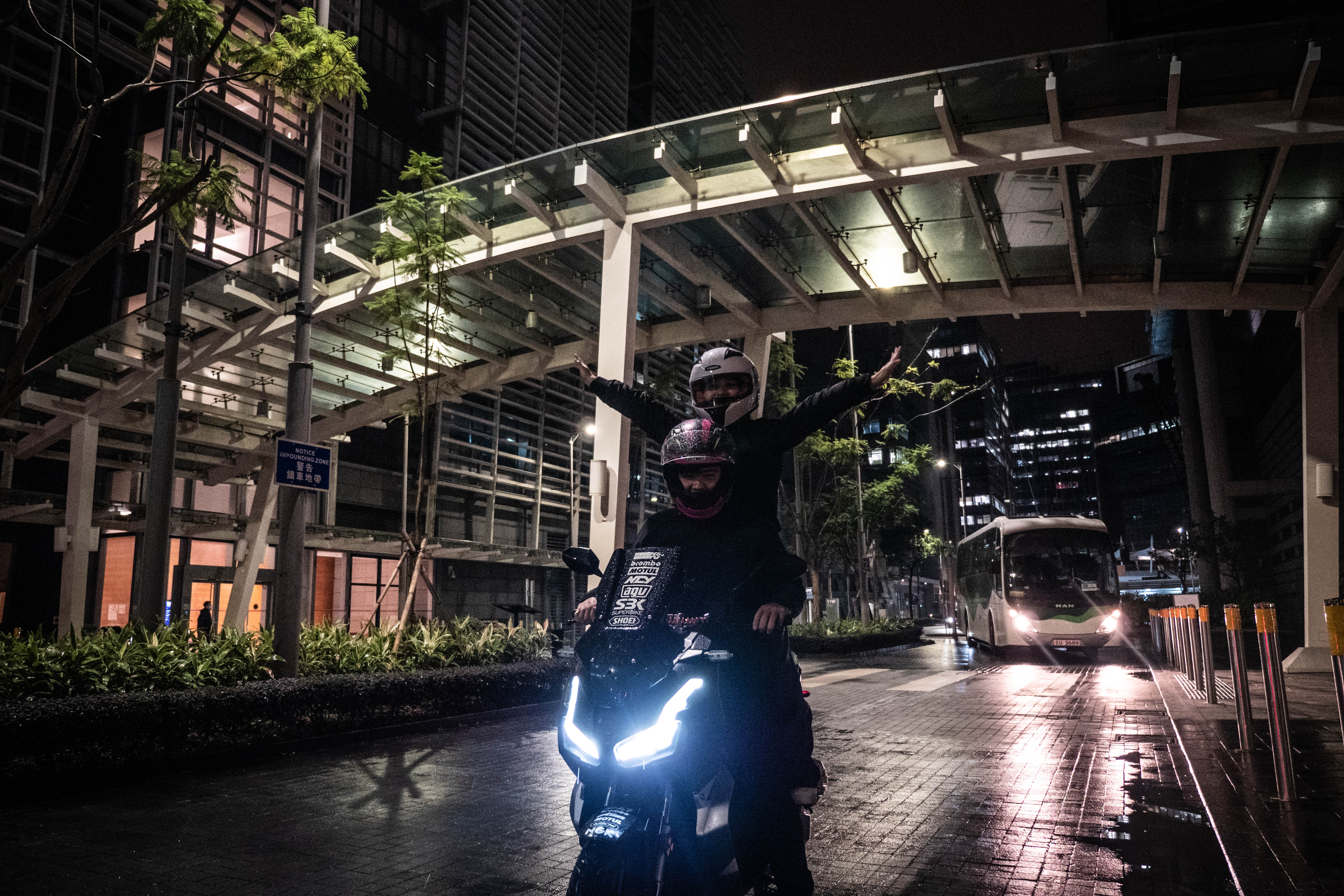 Two riders on the bike under the canopy, a bus passing on the wet street behind