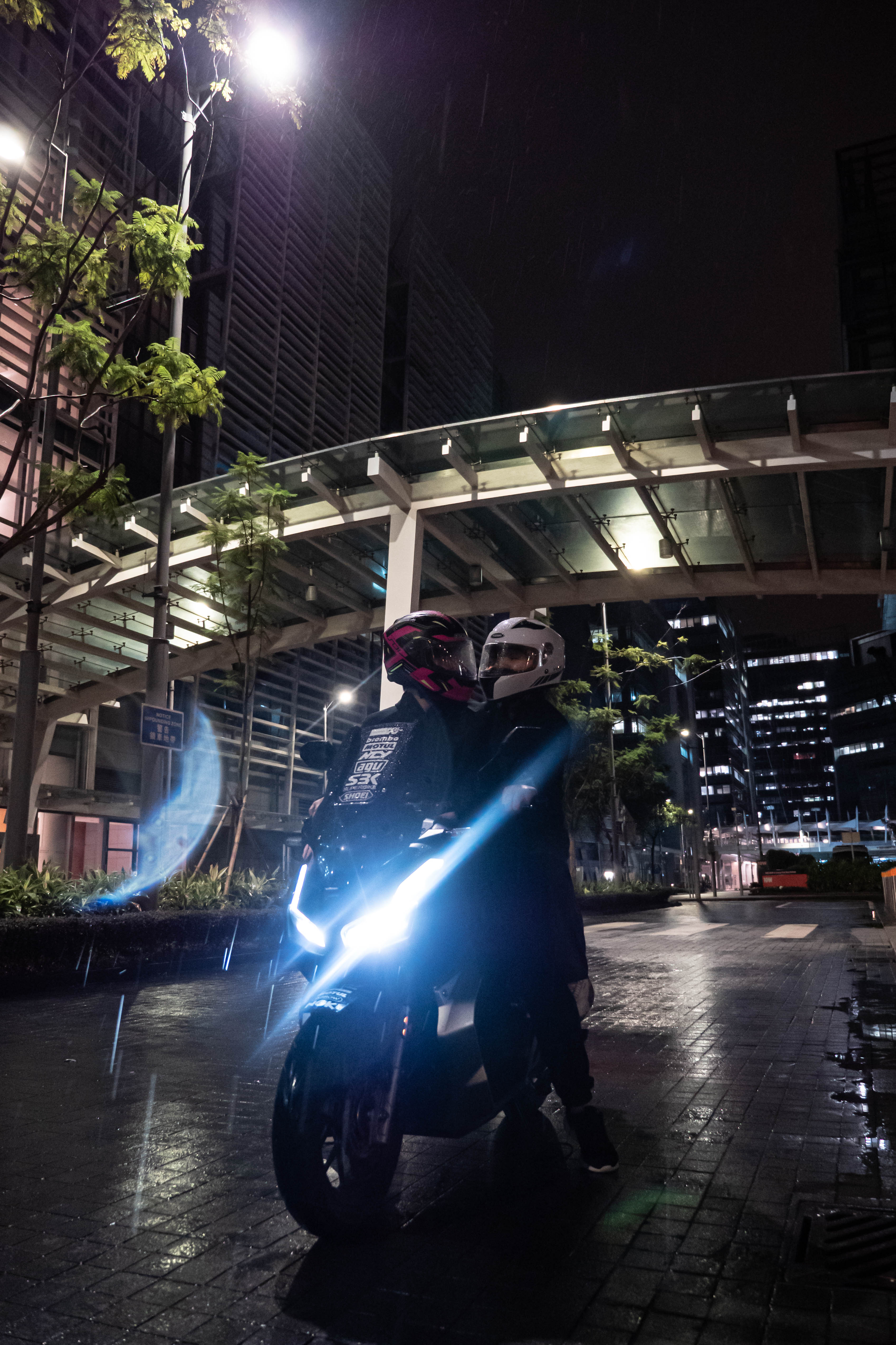 Two riders on a motorcycle under a lit canopy, the headlight glowing on wet paving