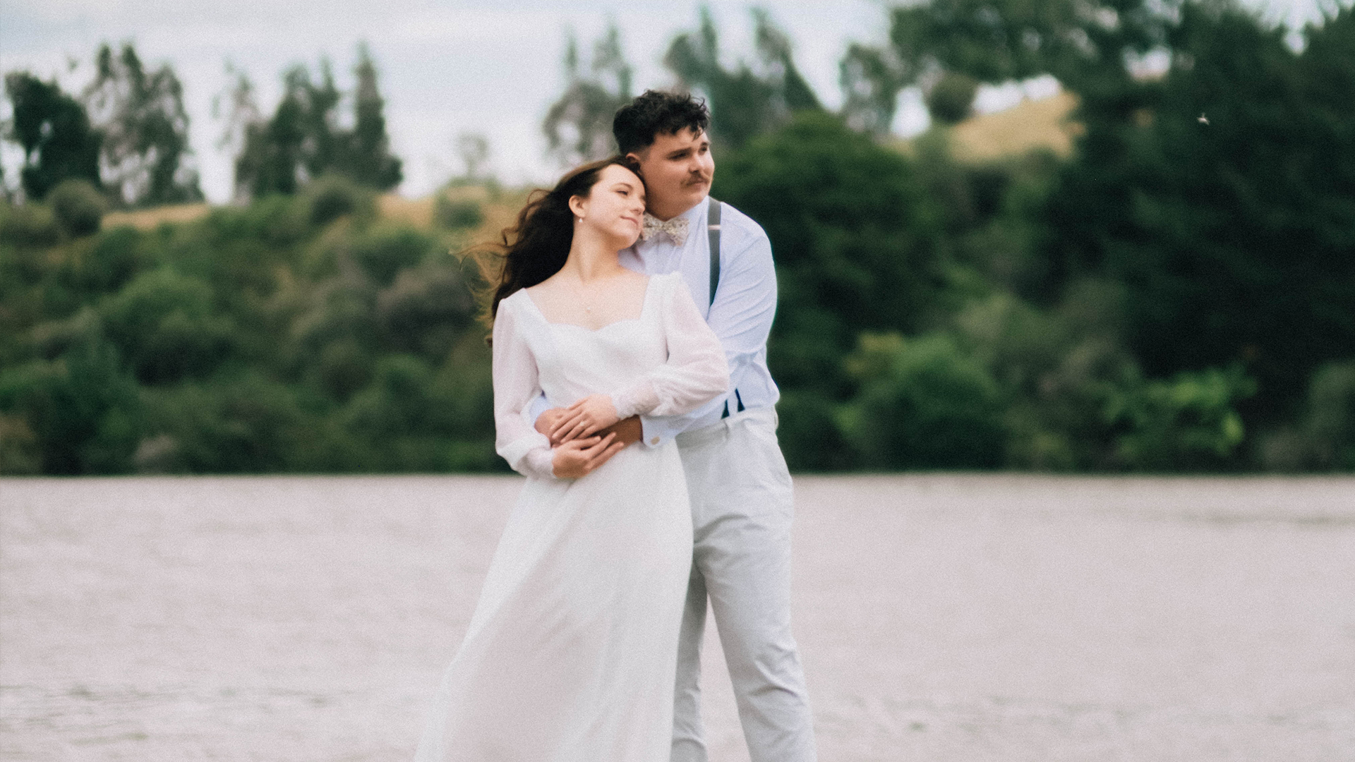 Joe hugging Mia from behind on a lakeshore, Mia in a long-sleeved lace wedding dress