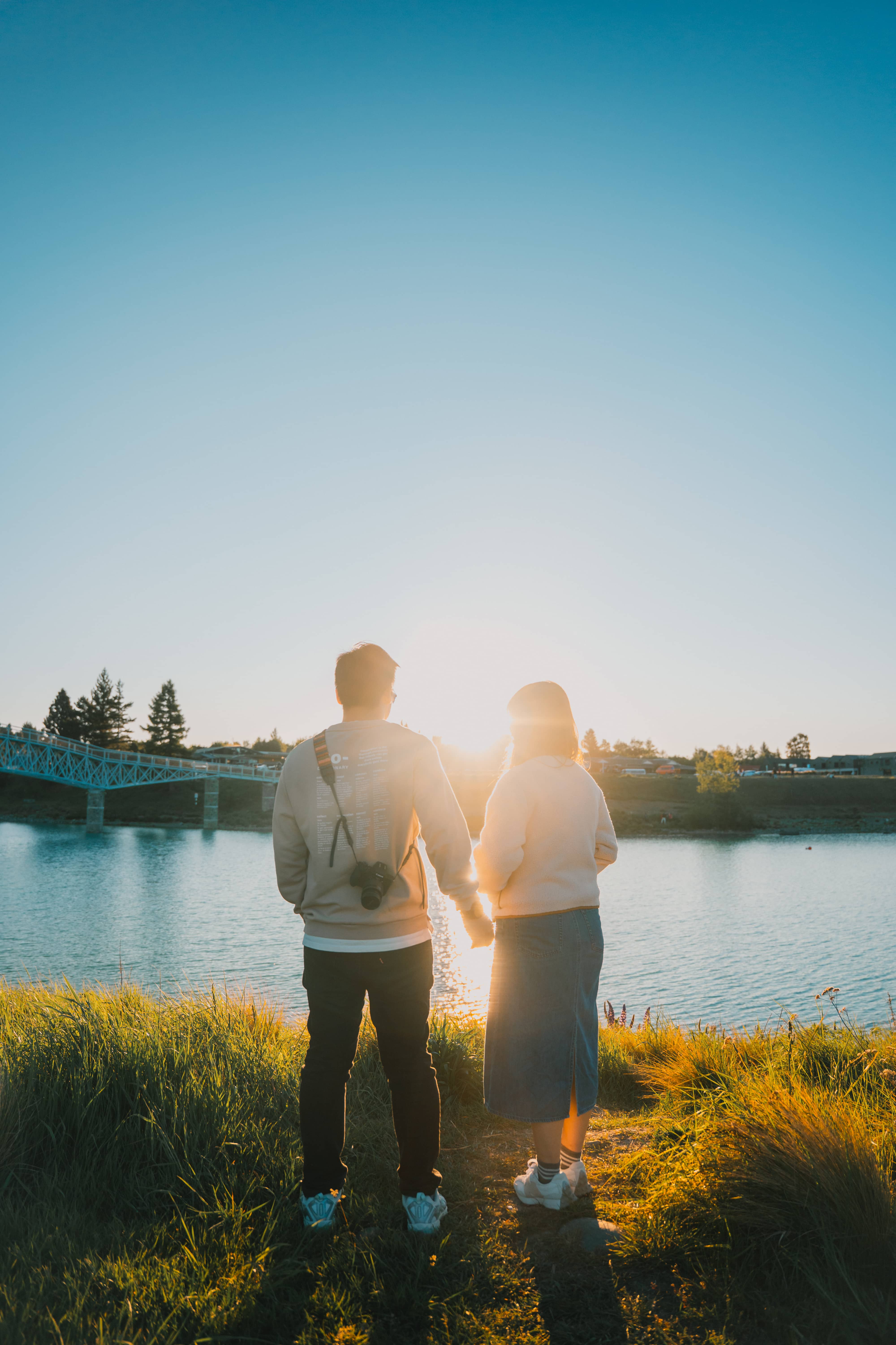 Julio and Kanis holding hands with their backs to the camera, looking across a lake at sunset