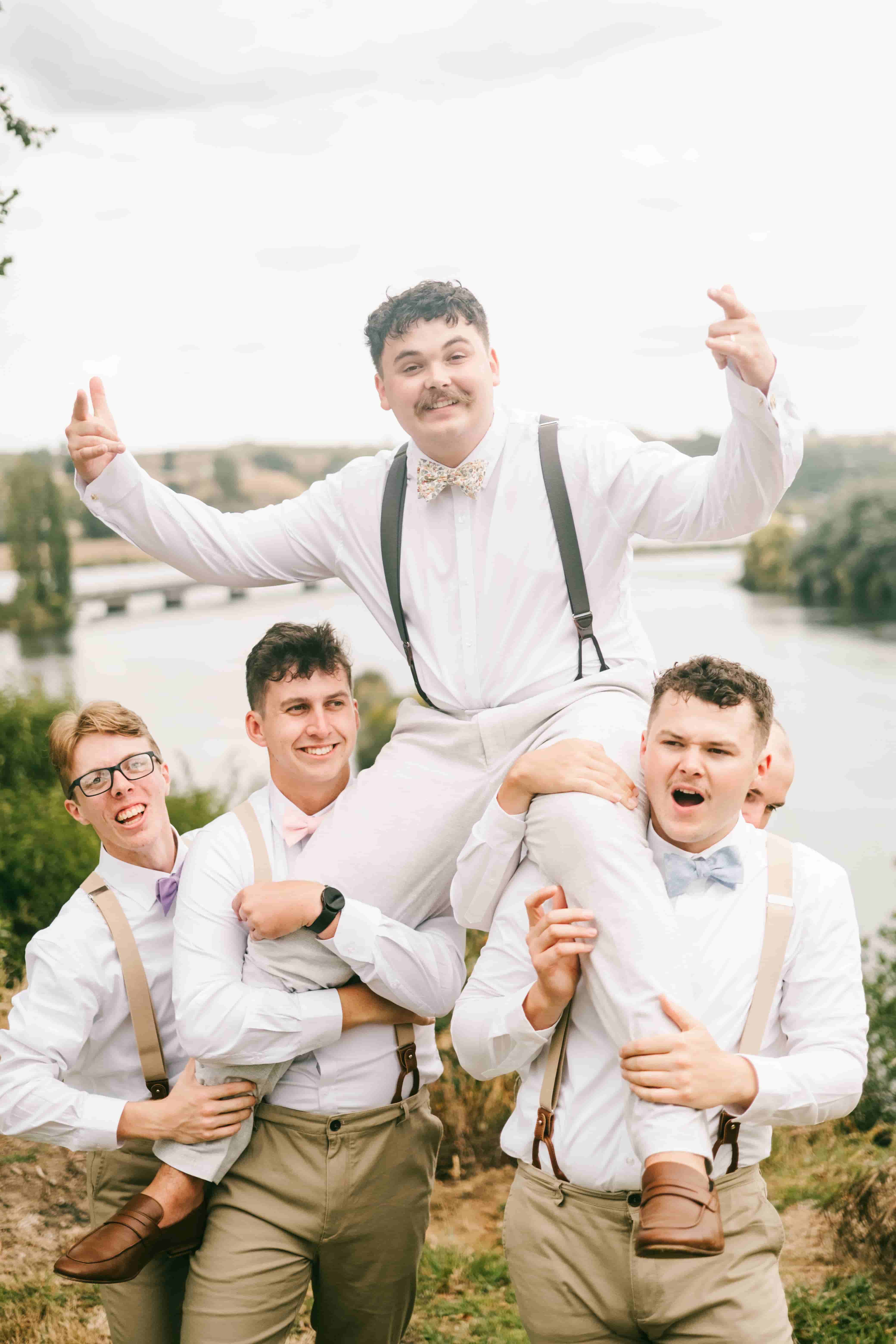 Groomsmen hoisting Joe onto their shoulders as he throws his arms out beside the water