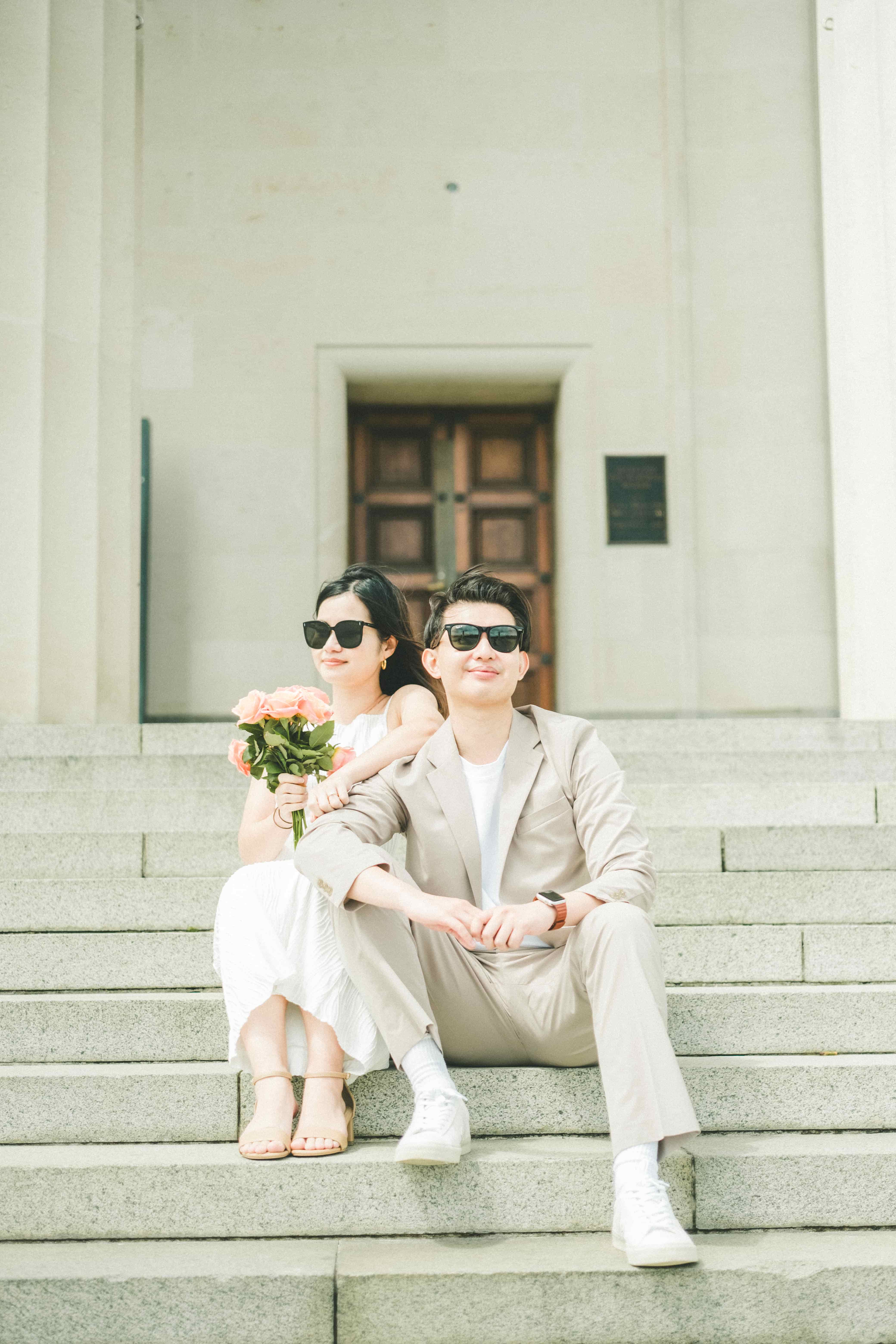 Suzen and Nick in sunglasses sitting on wide stone steps, Suzen holding a peach rose bouquet