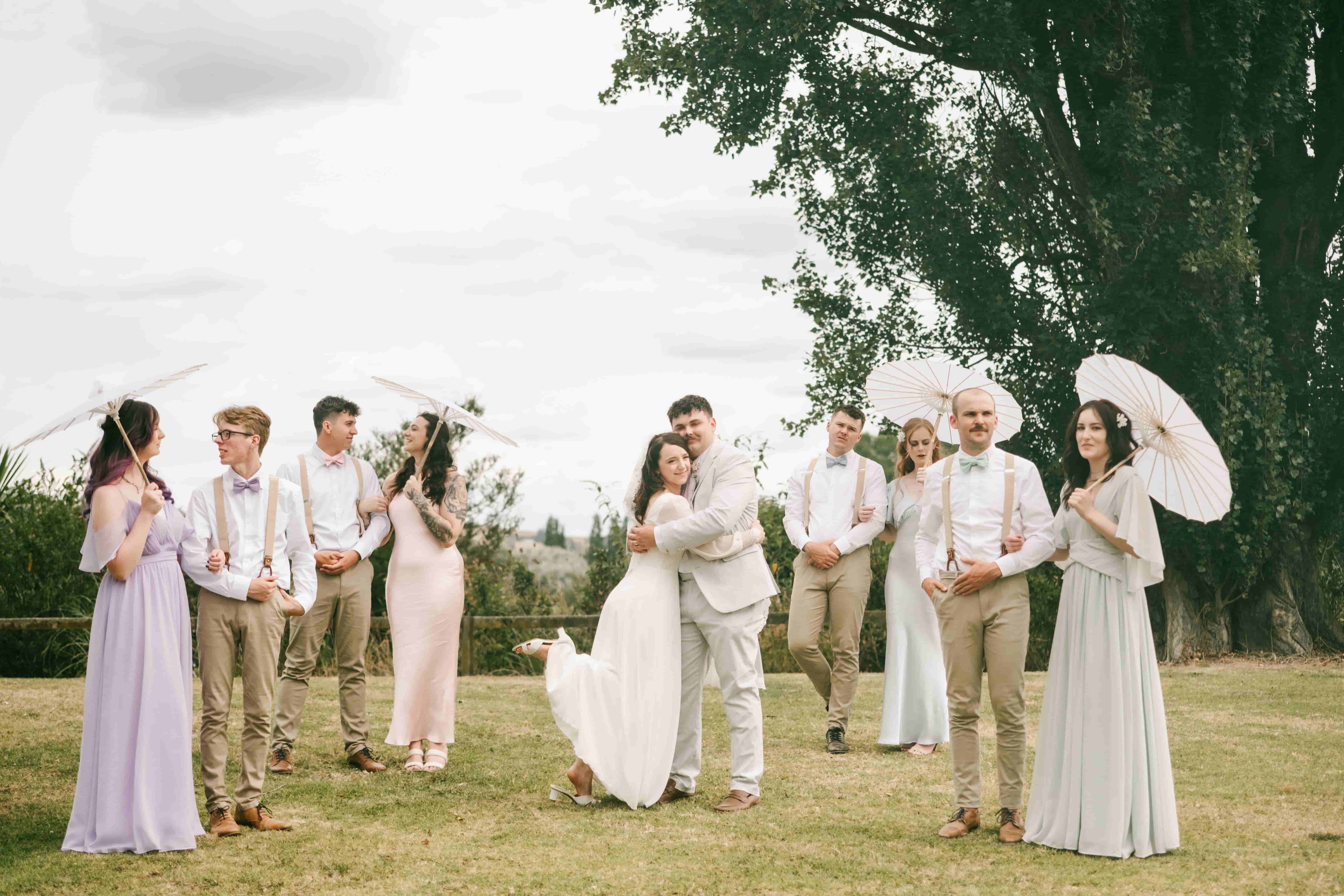Mia and Joe embracing in the middle of the wedding party on a grassy field under big trees