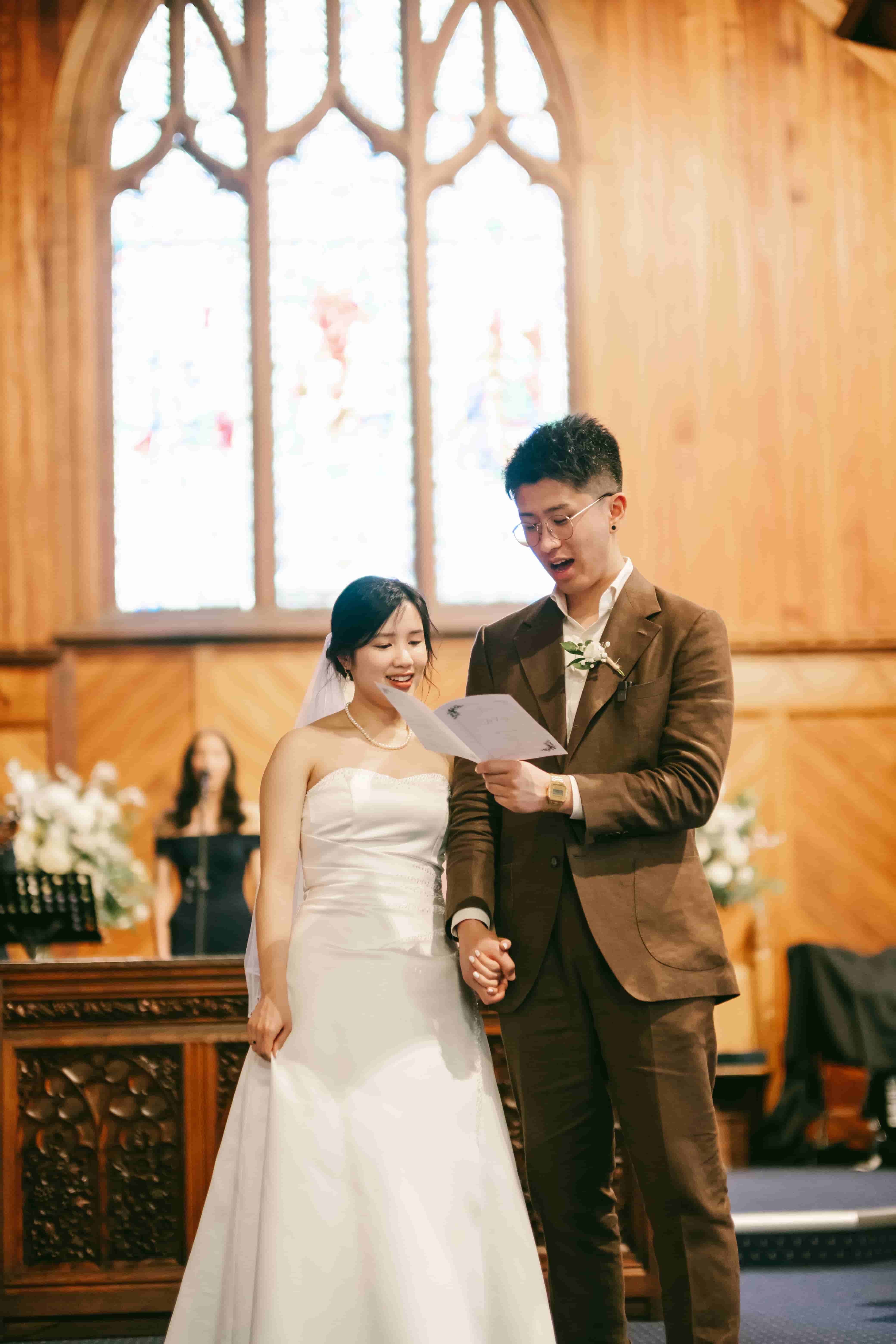 Sam reading his vows from a card as May smiles beside him, a stained glass window behind