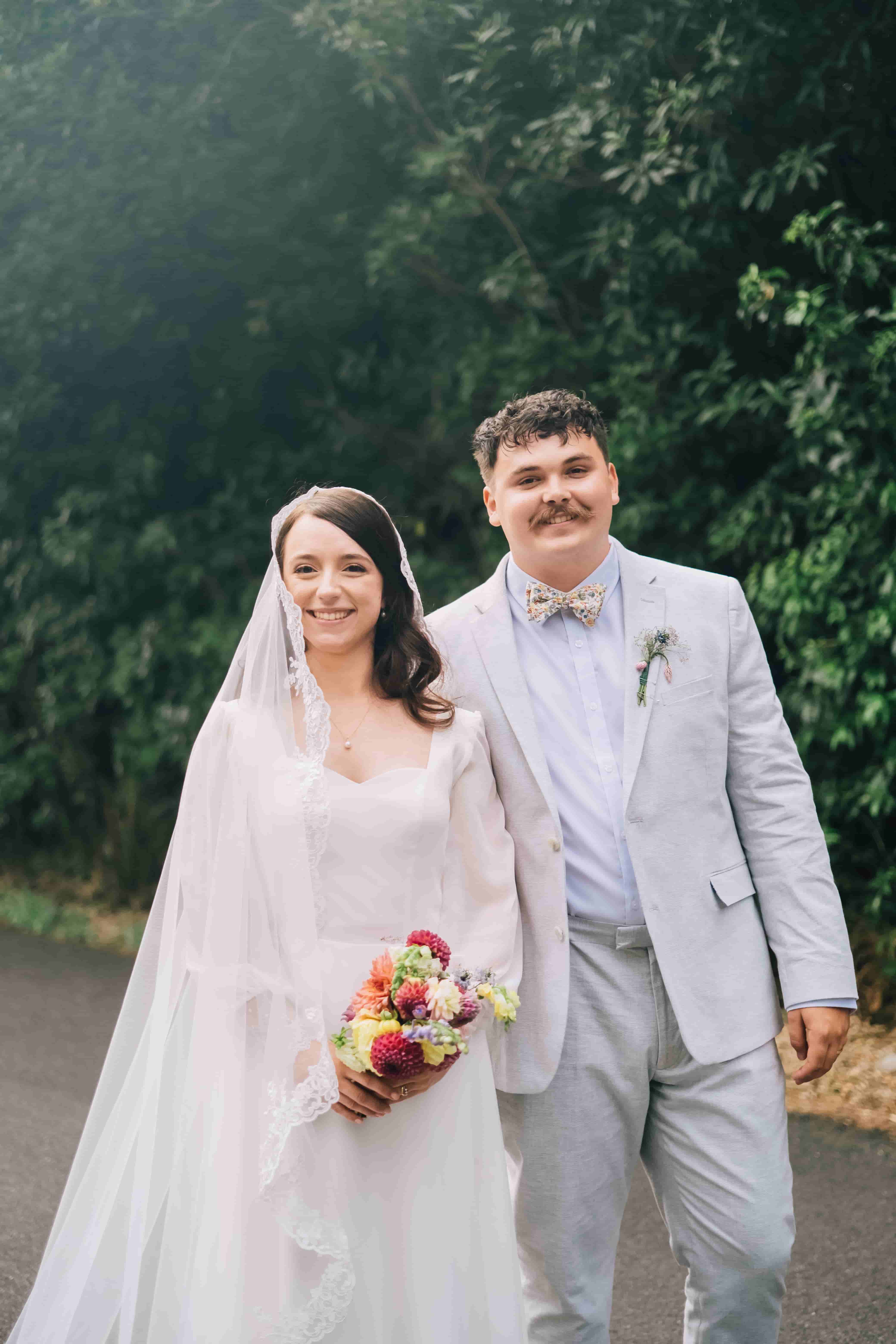 Mia in her lace veil and Joe in a pale grey suit smiling together on a tree-lined road
