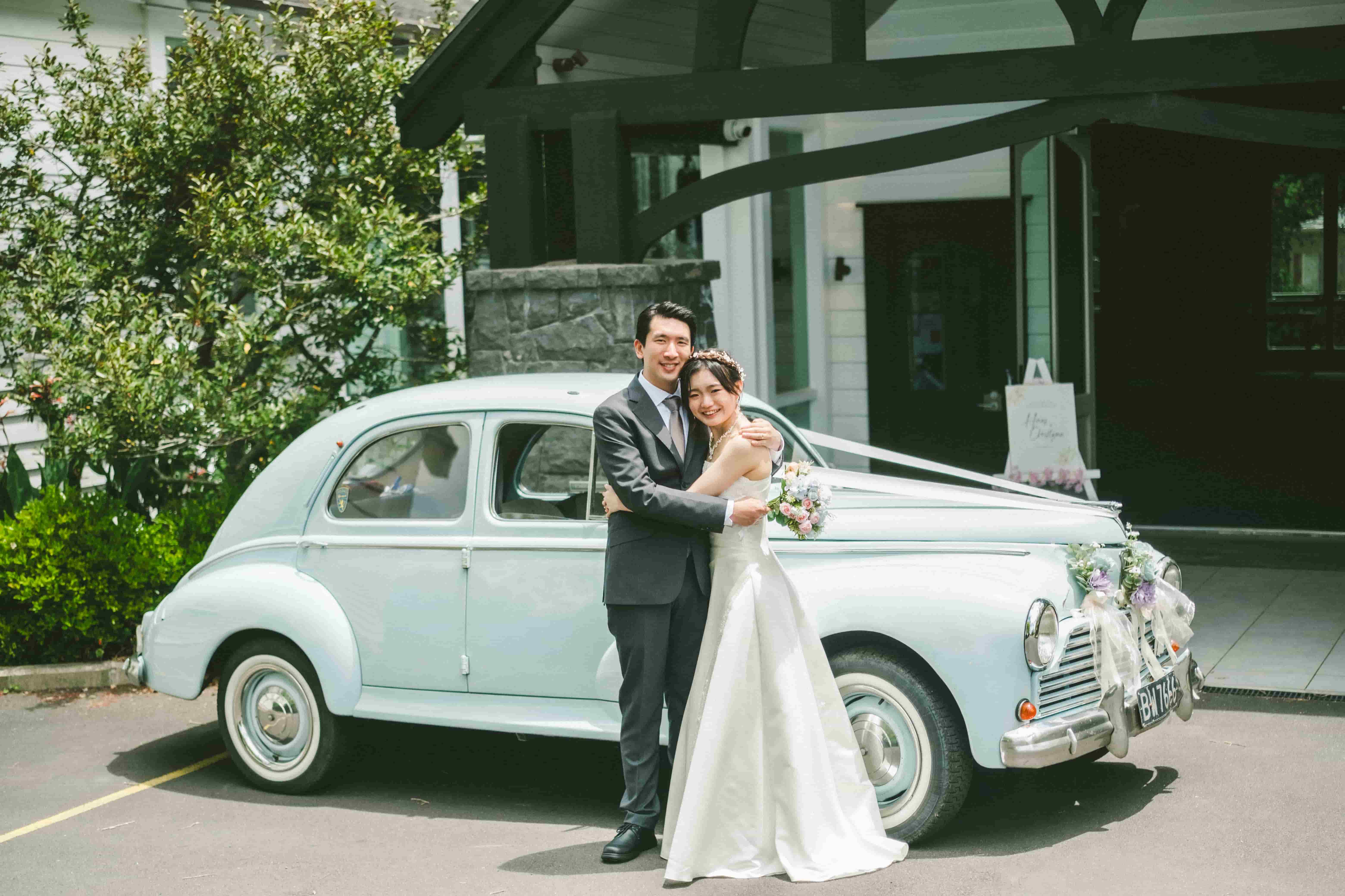 Hans and Chrystynn hugging beside a vintage wedding car outside the church